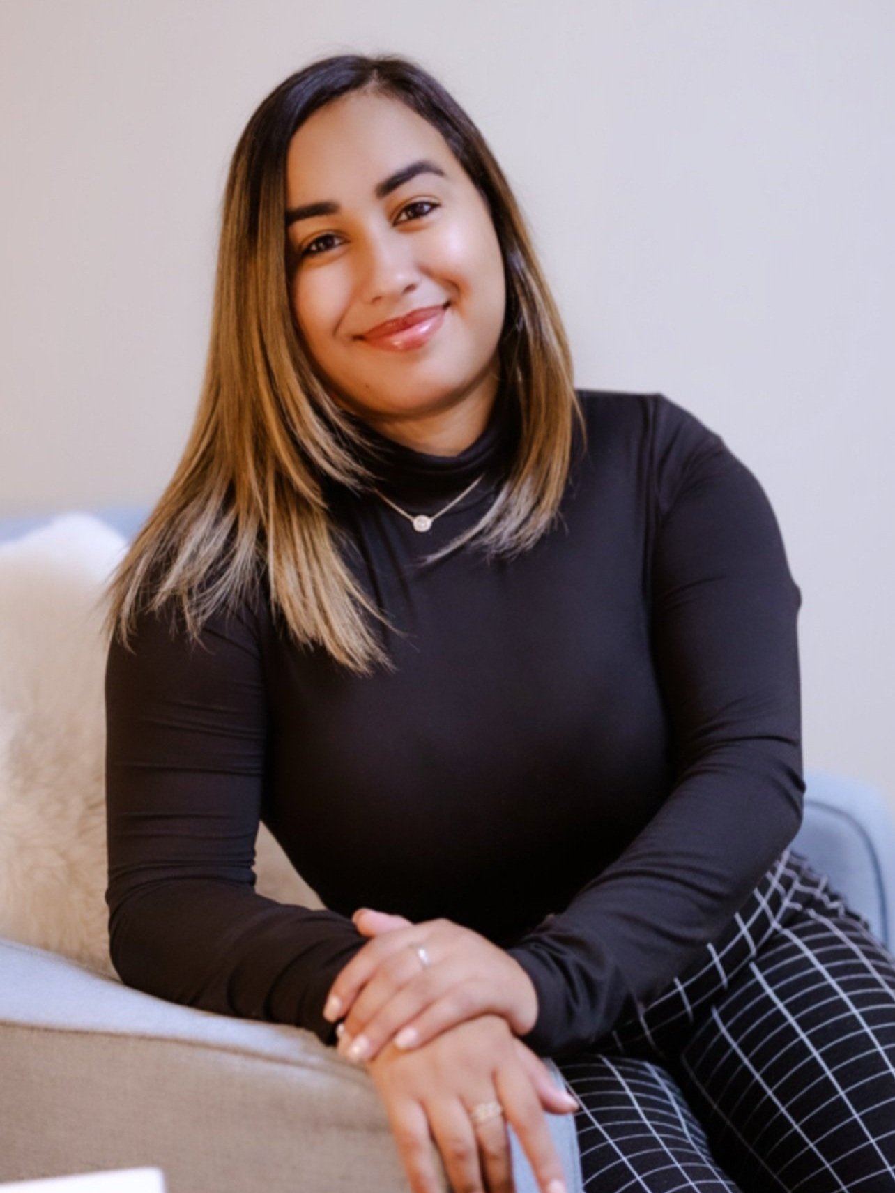 A young woman with shoulder-length straight hair, wearing a black turtleneck top, sitting on a couch, smiling, with her hands resting on her lap, against a plain light-colored background.