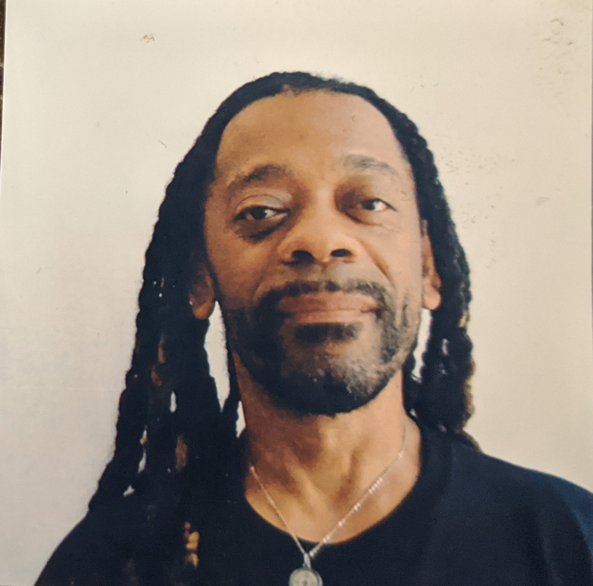 Portrait of a man with long dreadlocks, facial hair, wearing a black shirt and a silver necklace with a pendant, against a plain wall.