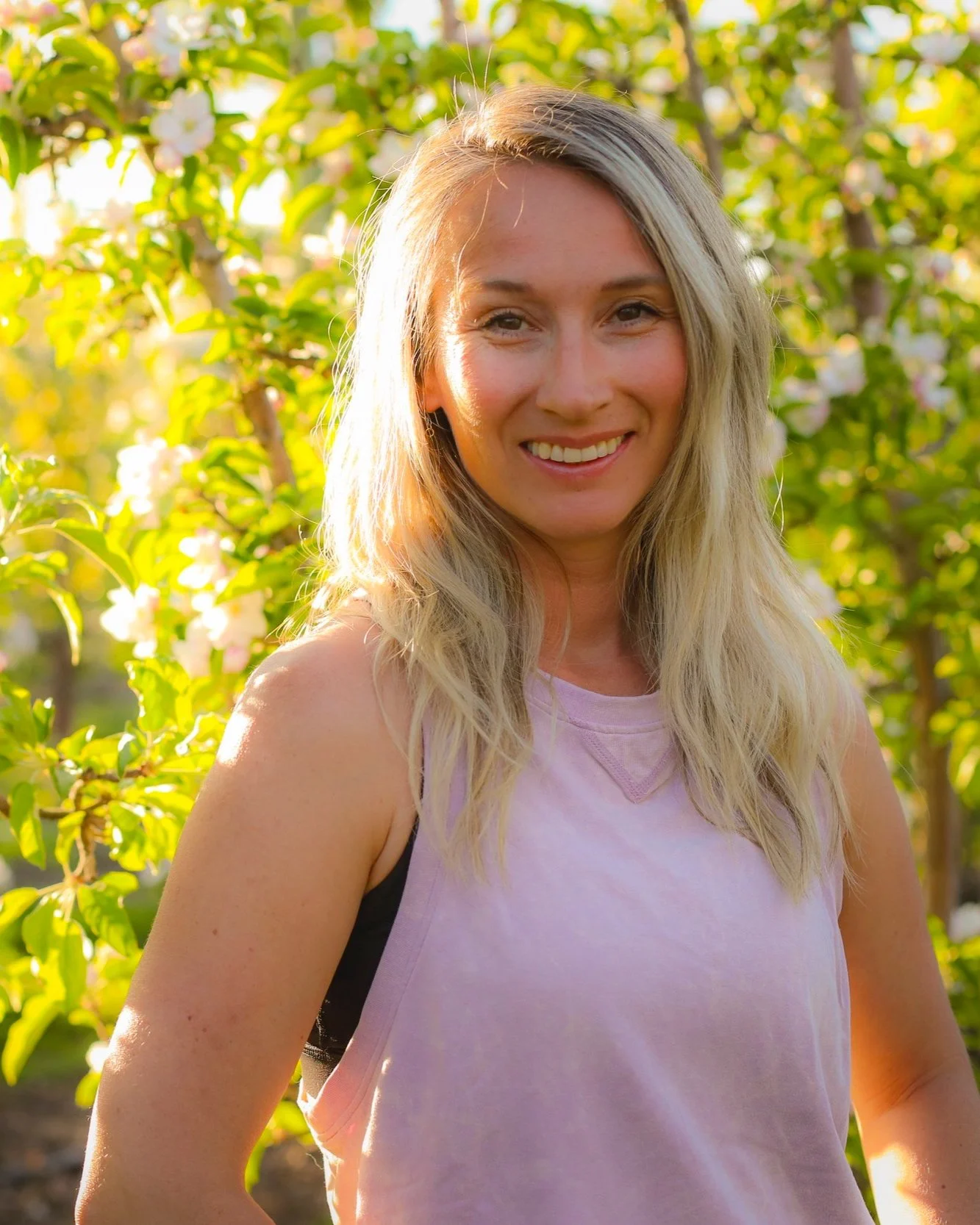 A smiling young woman with blonde hair standing outdoors in front of flowering bushes, illuminated by warm sunlight.