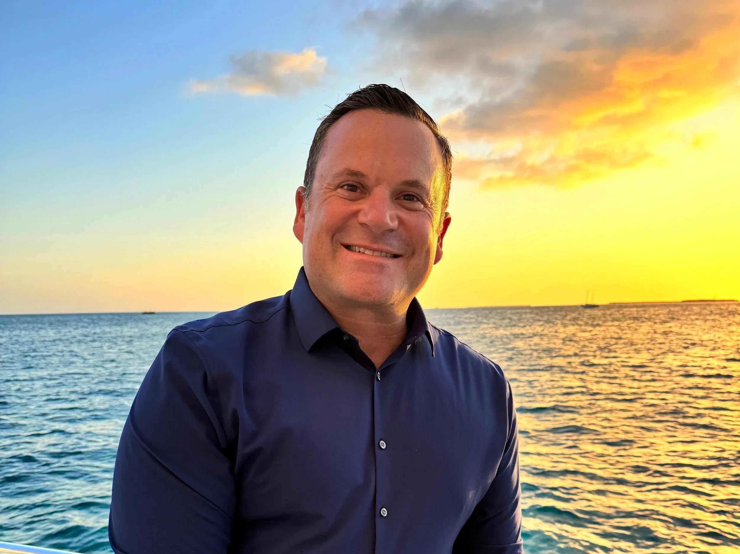 A man in a dark blue shirt smiling at sunset by the water with a colorful sky and the ocean behind him.