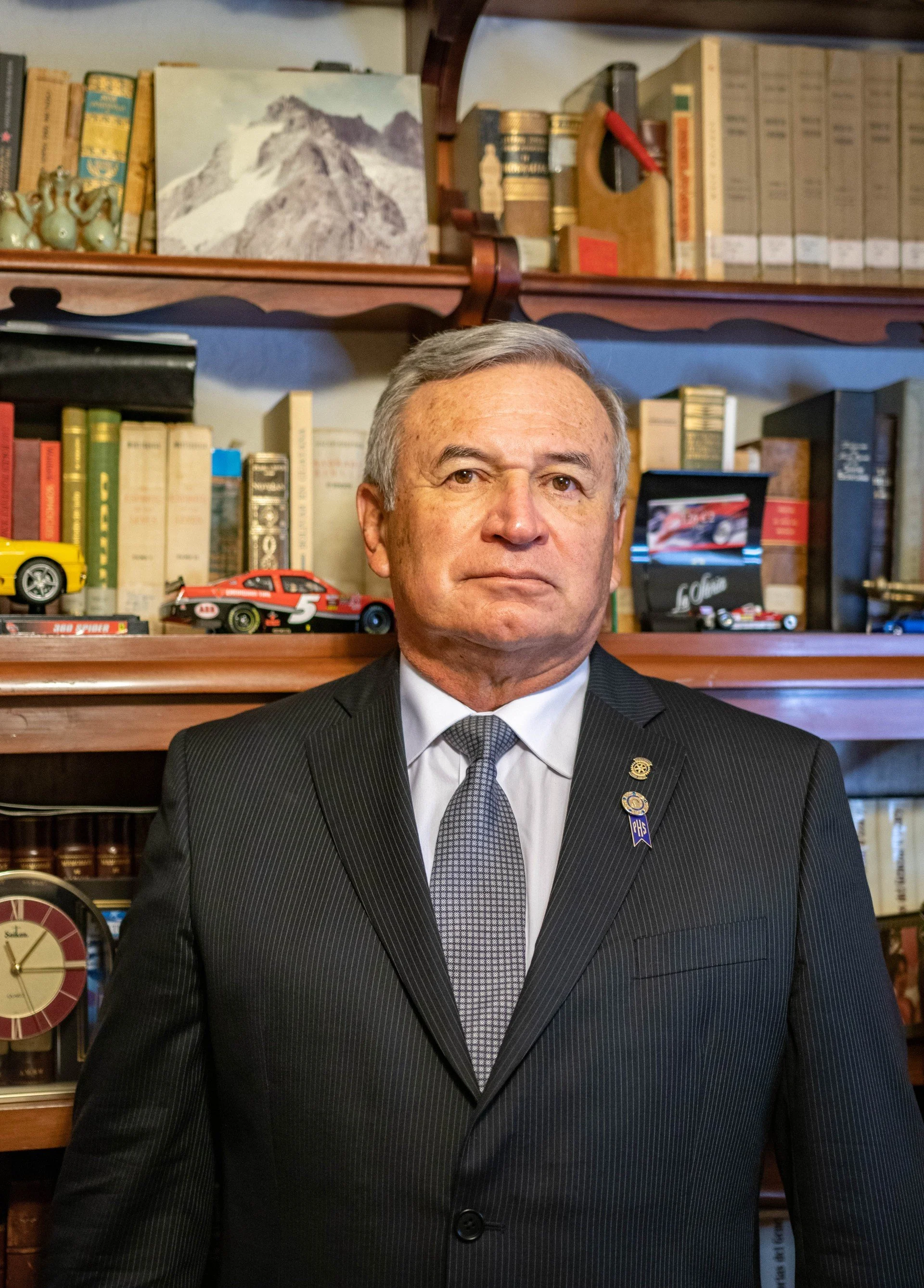 A middle-aged man in a dark pinstripe suit with a patterned tie and pins on his lapel, standing in front of a bookshelf with books, model cars, and decorative items.