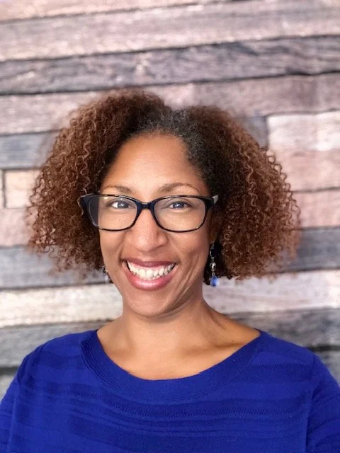 A woman with curly hair, glasses, and earrings smiling in front of a wooden background.