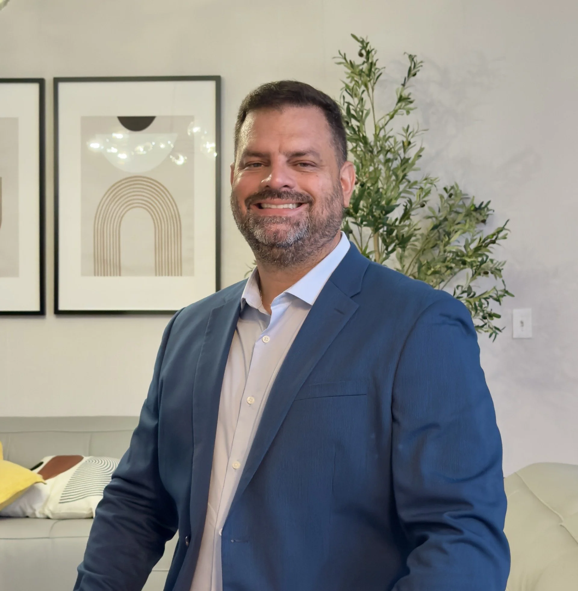 A smiling man with dark hair and a beard, wearing a blue suit and white shirt, standing indoors with a sofa, framed abstract art, and a tall plant in the background.