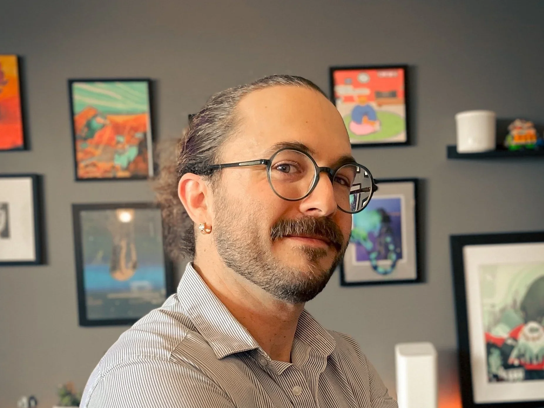 A man with glasses, a beard, and an earring smiling at the camera inside an art-filled room.