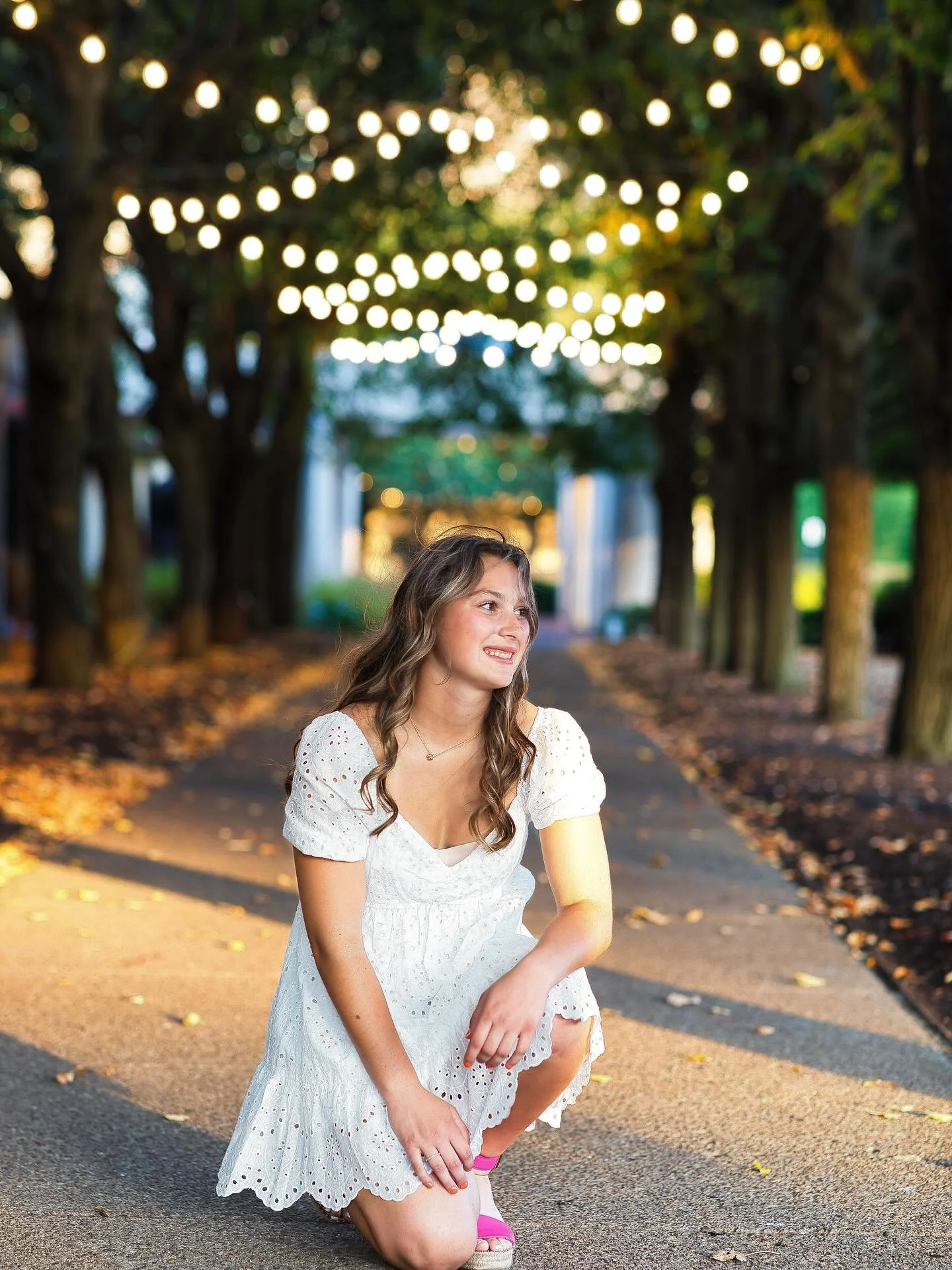 Senior girl in white dress posed on tree-lined walkway with warm string lights bokeh, Memorial Art Gallery Rochester NY senior portraits