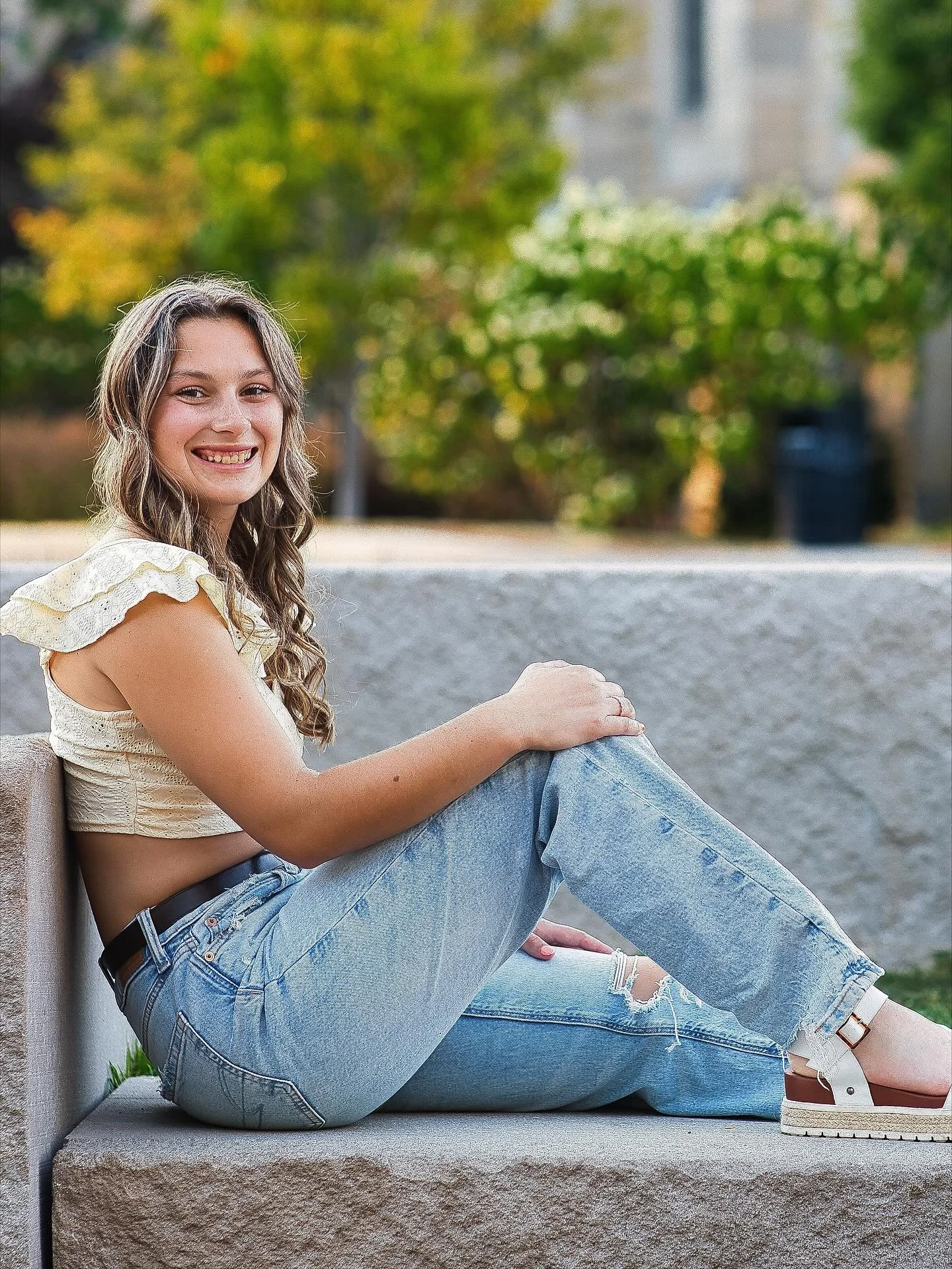Senior girl in white lace crop top and jeans sitting on stone ledge smiling, Memorial Art Gallery Rochester NY senior portraits