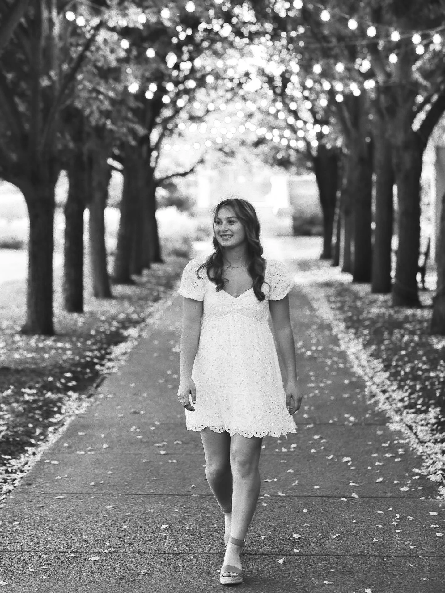 Black and white senior portrait of girl in white dress walking tree-lined path with string lights, Memorial Art Gallery Rochester NY