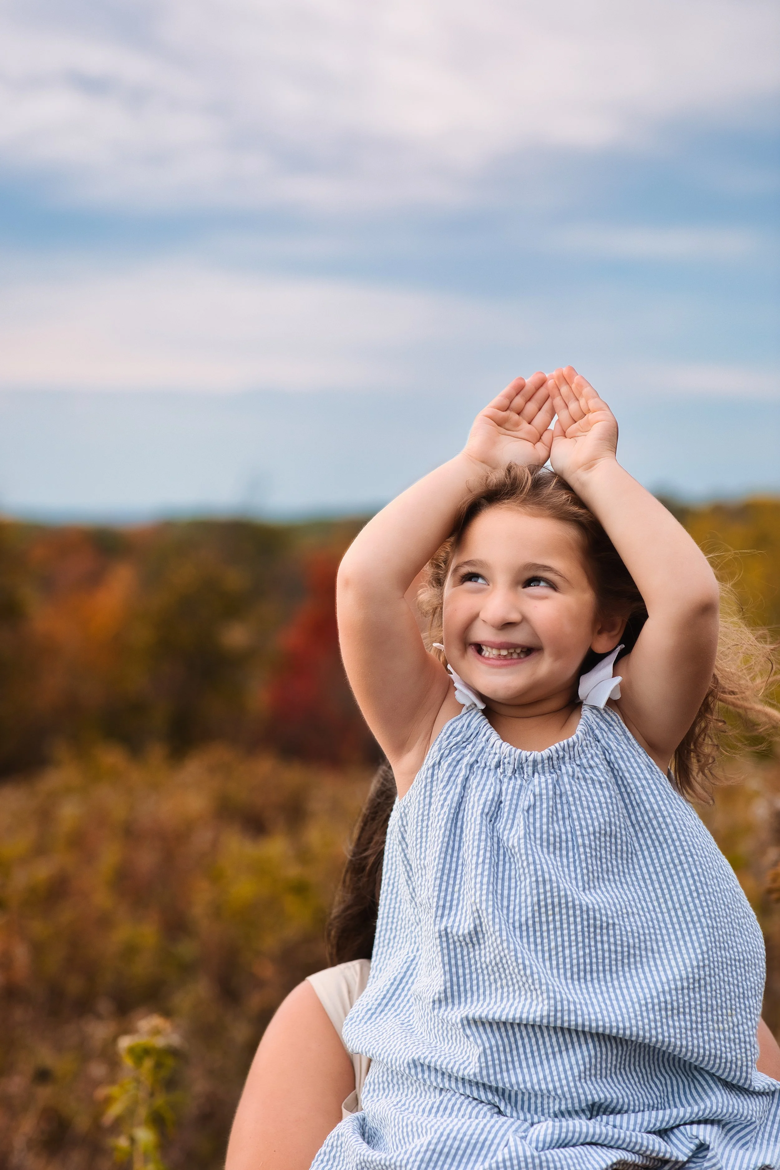Rochester, NY portrait of a young girl raising her hands and smiling outdoors with fall foliage