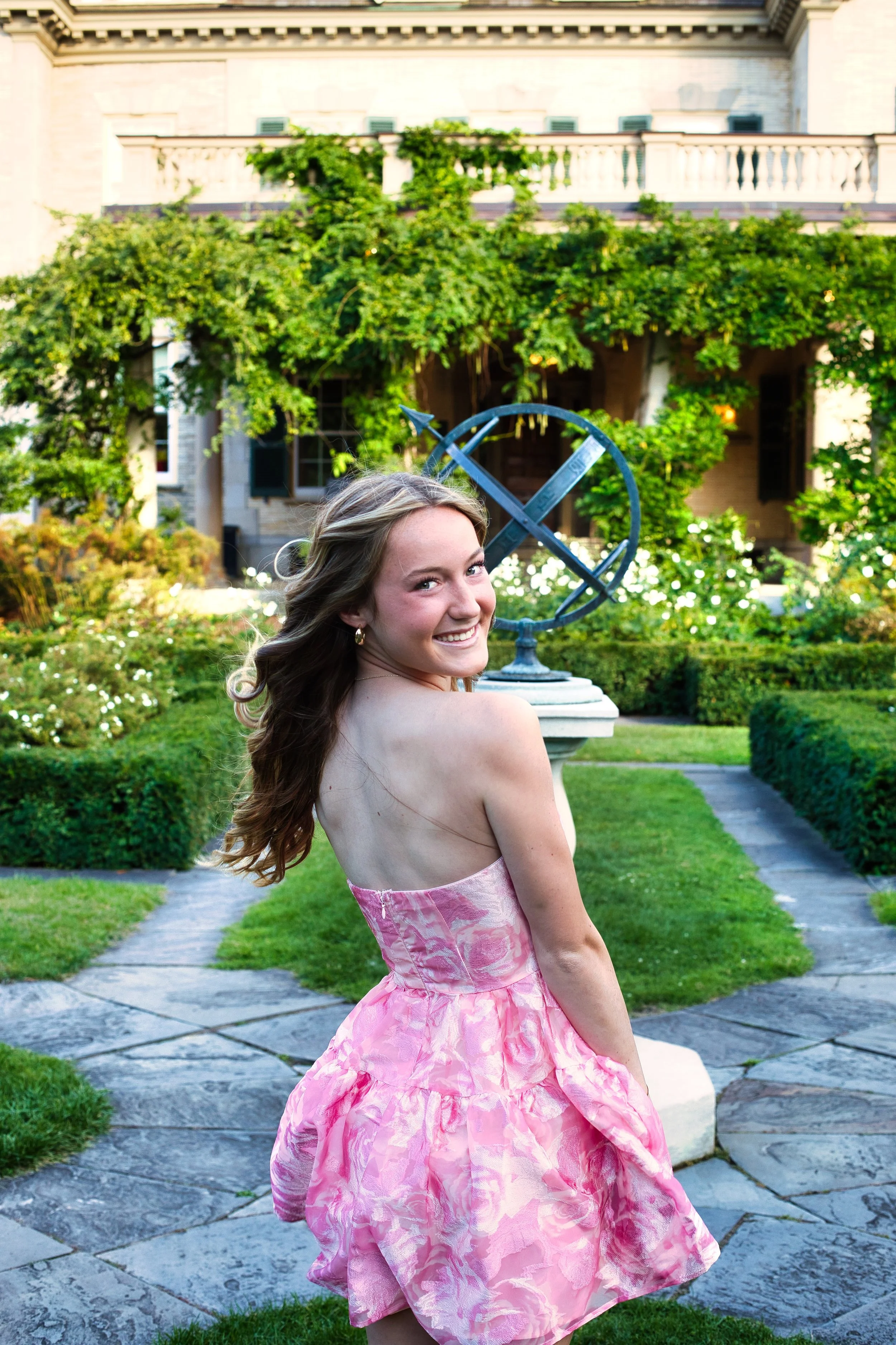 Senior girl in pink formal gown looking back over shoulder in manicured garden, George Eastman House Rochester NY senior pictures