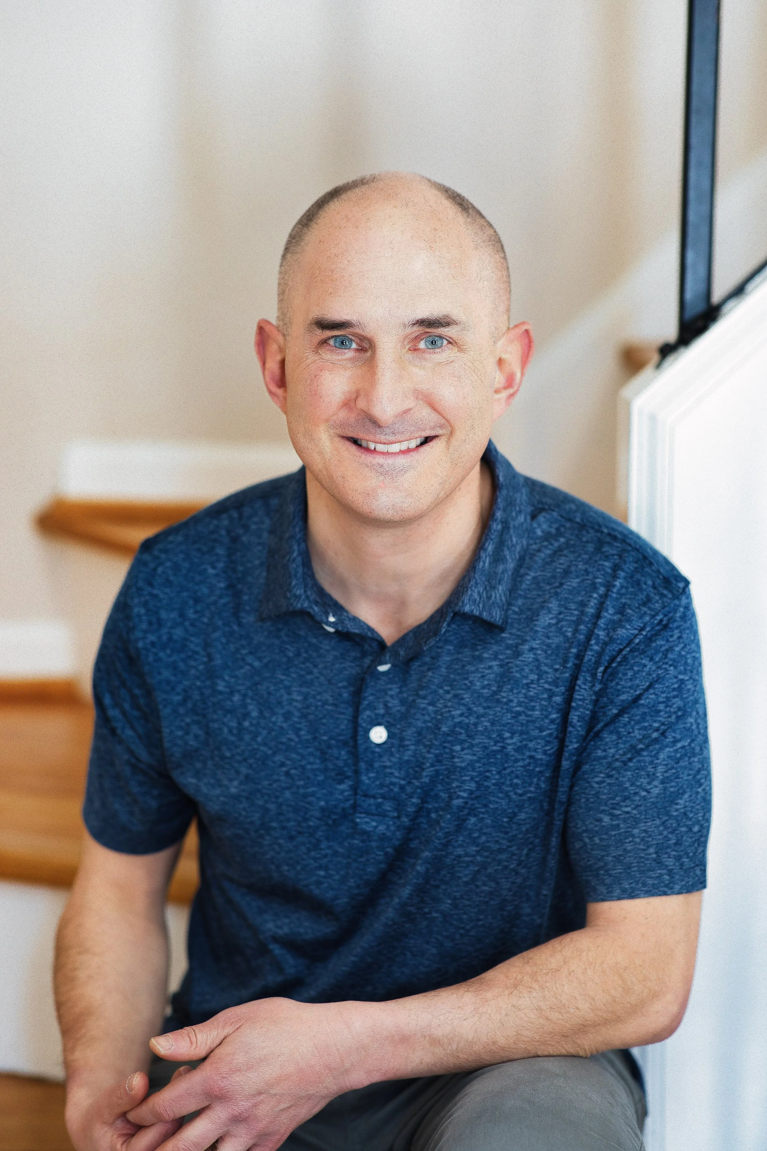 Relaxed business headshot of man in blue polo seated on stairs with natural light