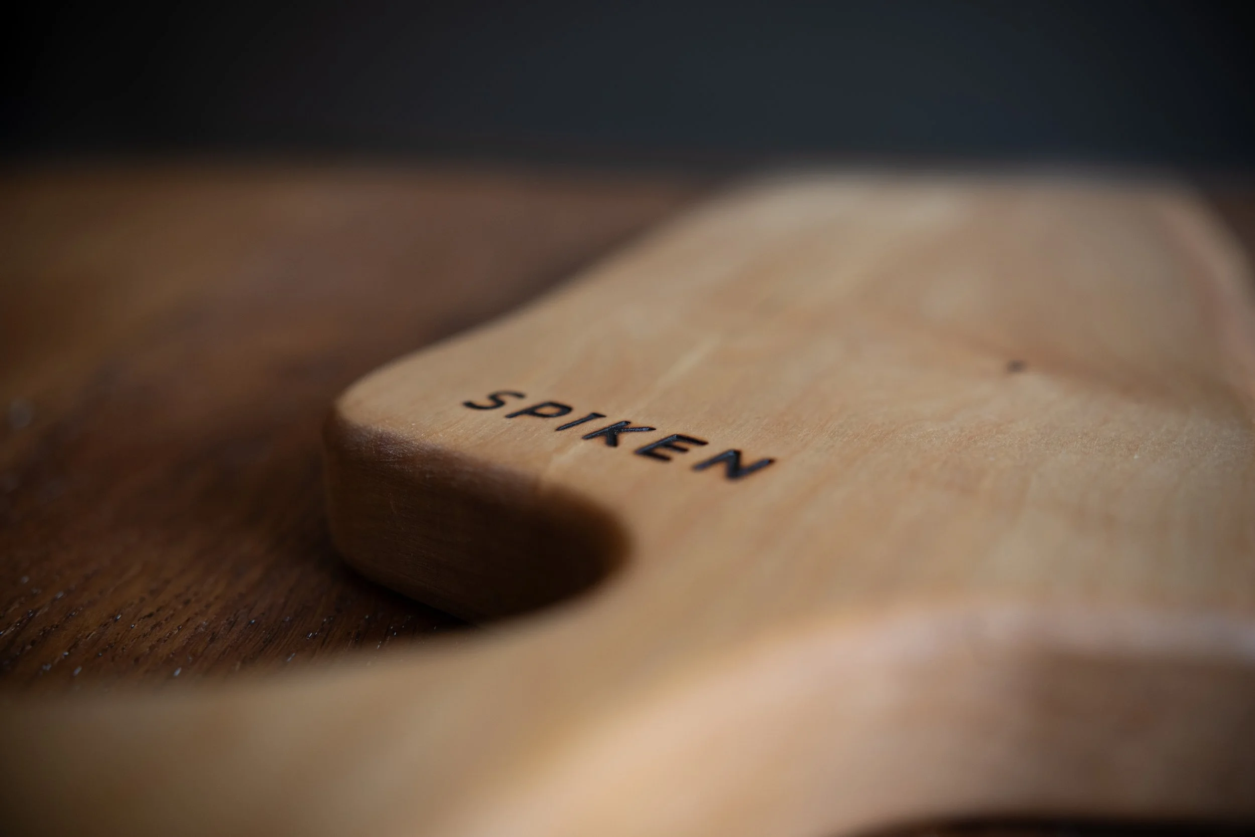 Ett träföremål med texten 'spiken' inristat på ytan, placerat på en träskiva. Handgjord serveringsbricka i massivt trä med unik ådring och naturlig finish. En stilren träprodukt som passar både vardag och fest.