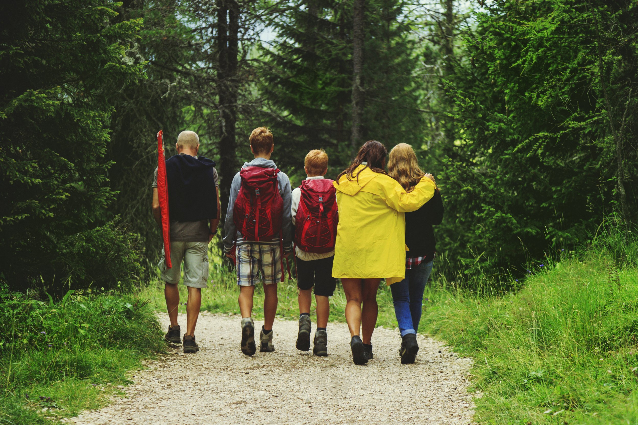 Six people, two adults and four children, walking on a forest trail with trees and greenery around them, some carrying backpacks and one carrying a red sleeping pad.
