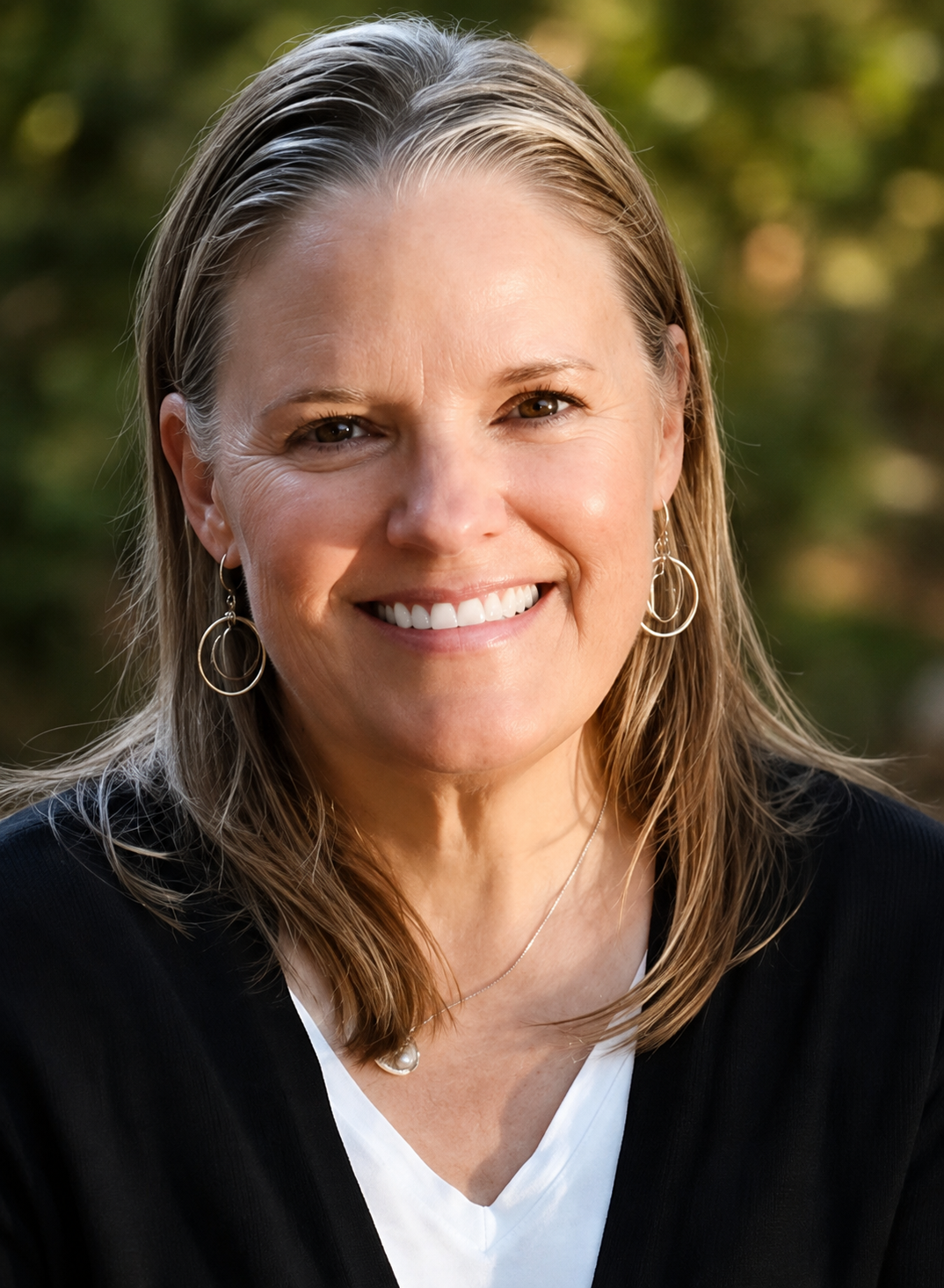 A smiling middle-aged woman with shoulder-length blonde hair, wearing earrings and a pearl necklace, outdoors with a blurred background of trees, in natural lighting.