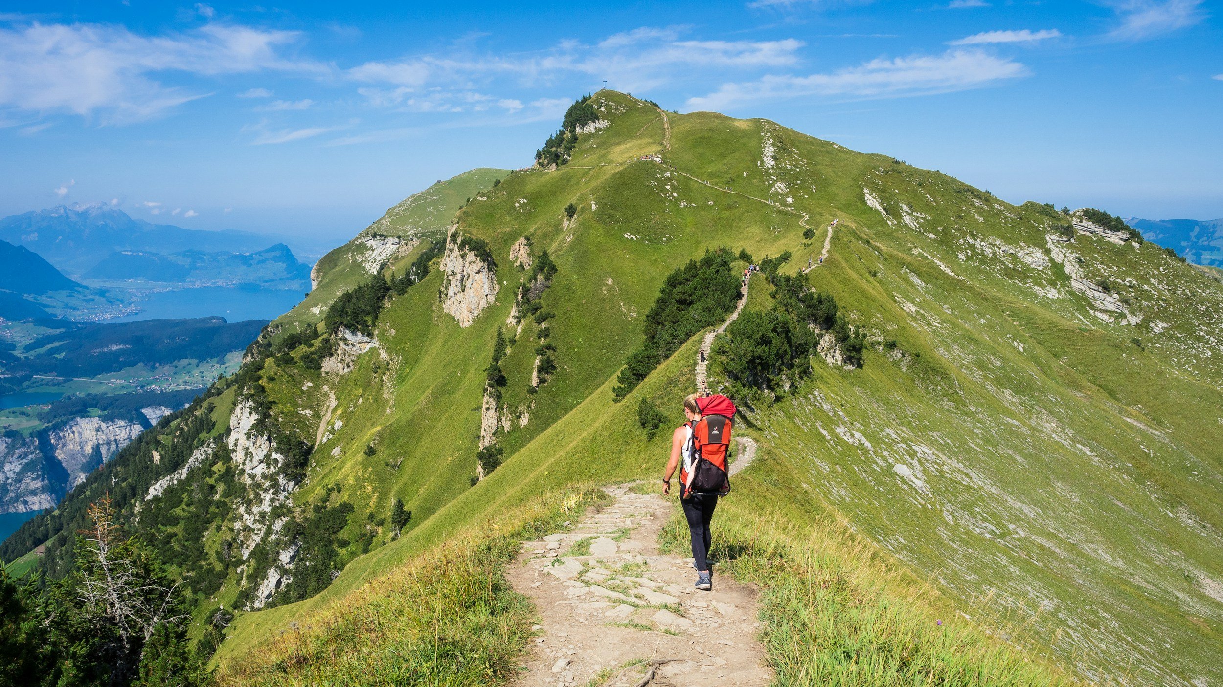 Hiker walking on a mountain trail with a backpack, green grassy slopes, and a scenic mountain landscape under a blue sky.