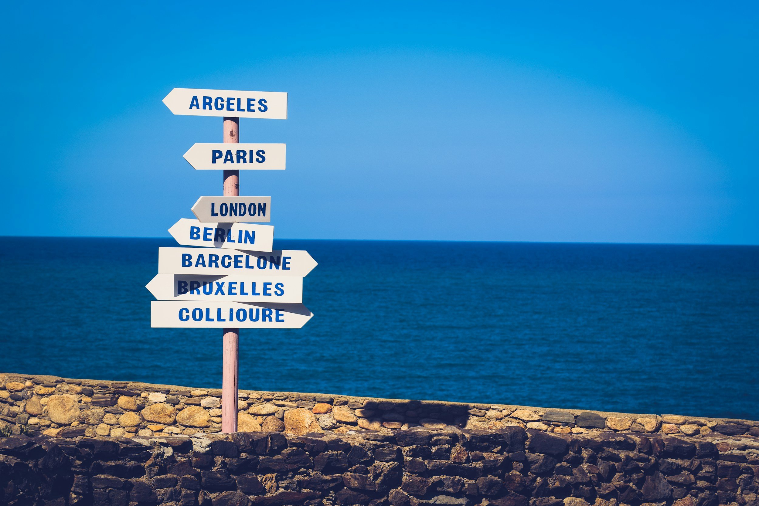 A white signpost with blue text and arrows pointing in different directions, located on a stone wall by the sea. Directions include Argelès, Paris, London, Berlin, Barcelone, Bruxelles, and Collioure.