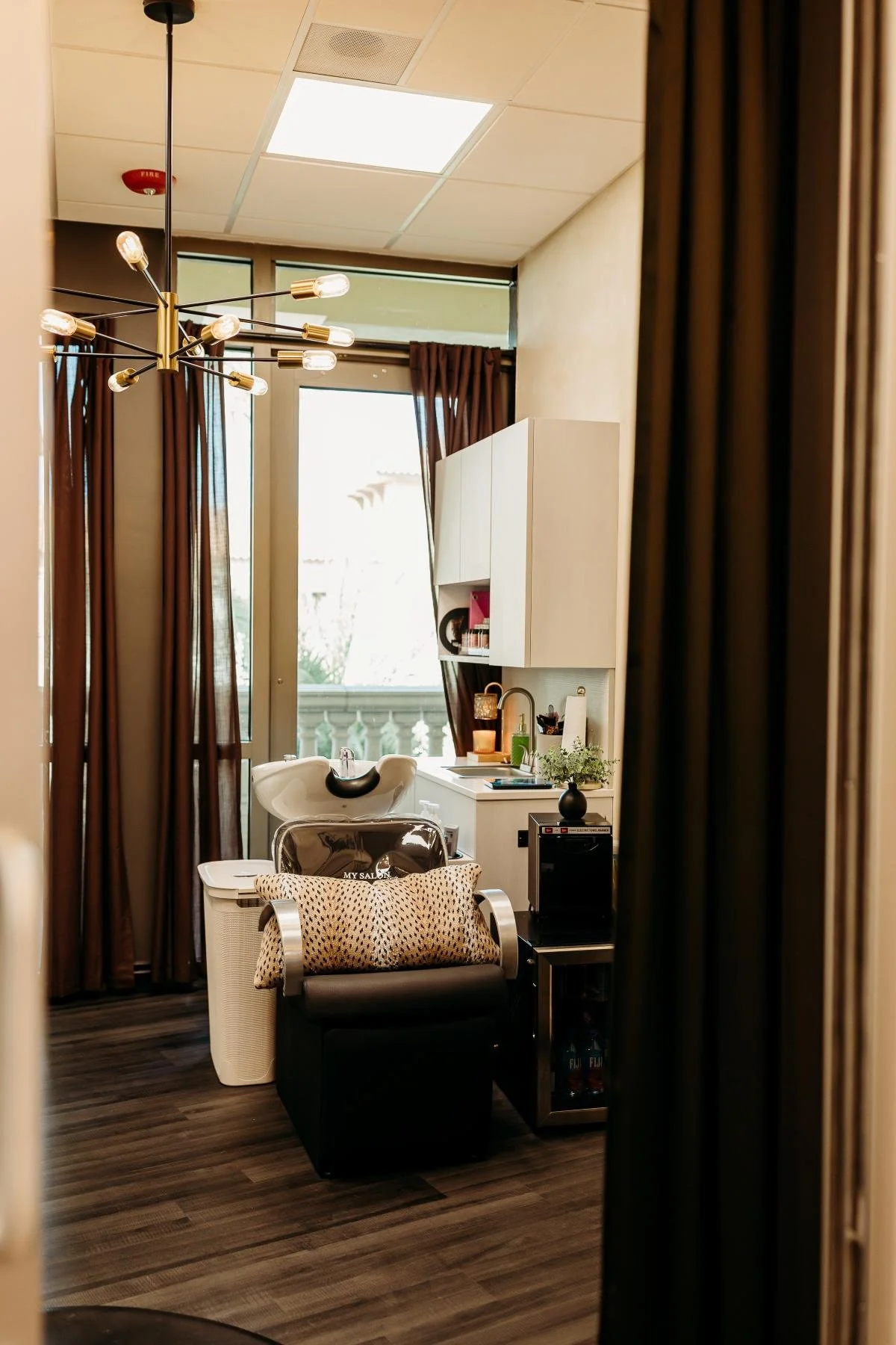A salon room with a hair washing station near a window, brown curtains, a black salon chair with a leopard print pillow, a small black refrigerator, and a countertop with hair and beauty products.