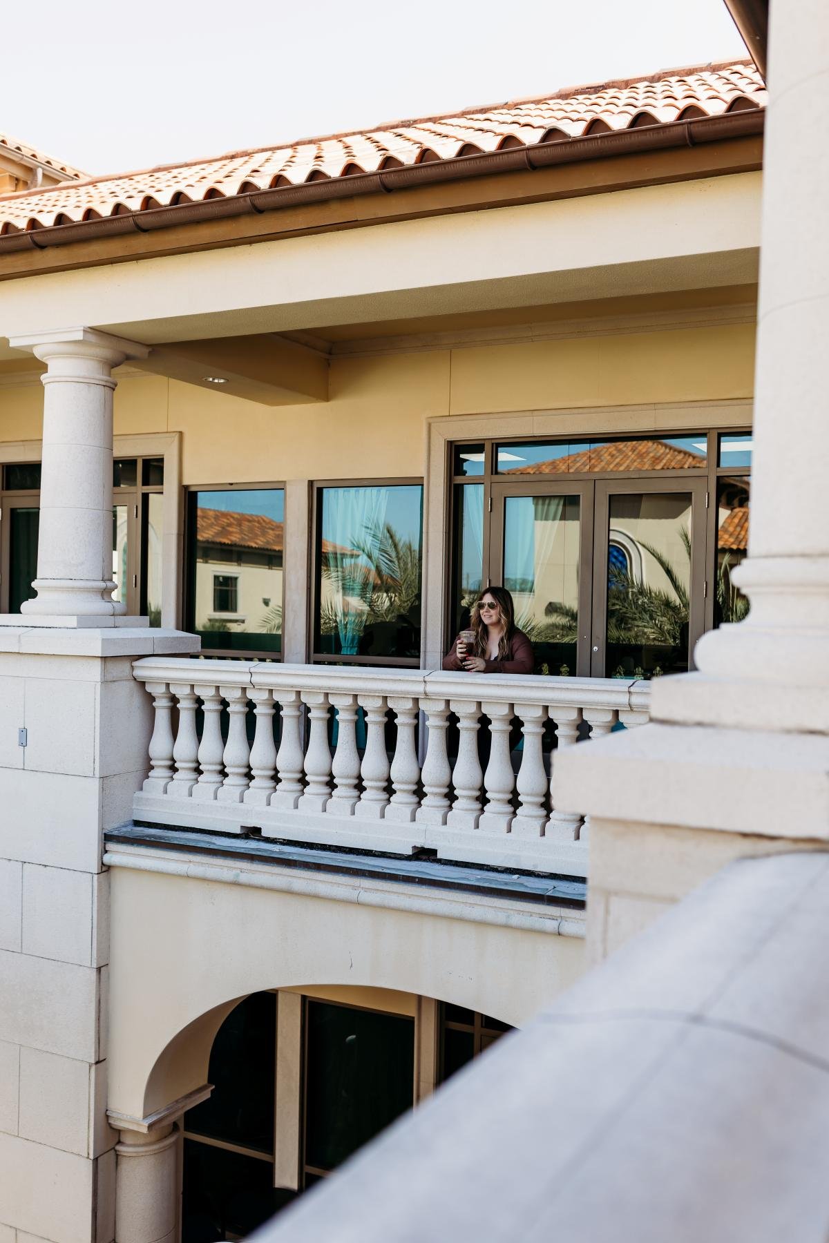 A woman with sunglasses sitting on a balcony of a house, holding a drink and smiling.