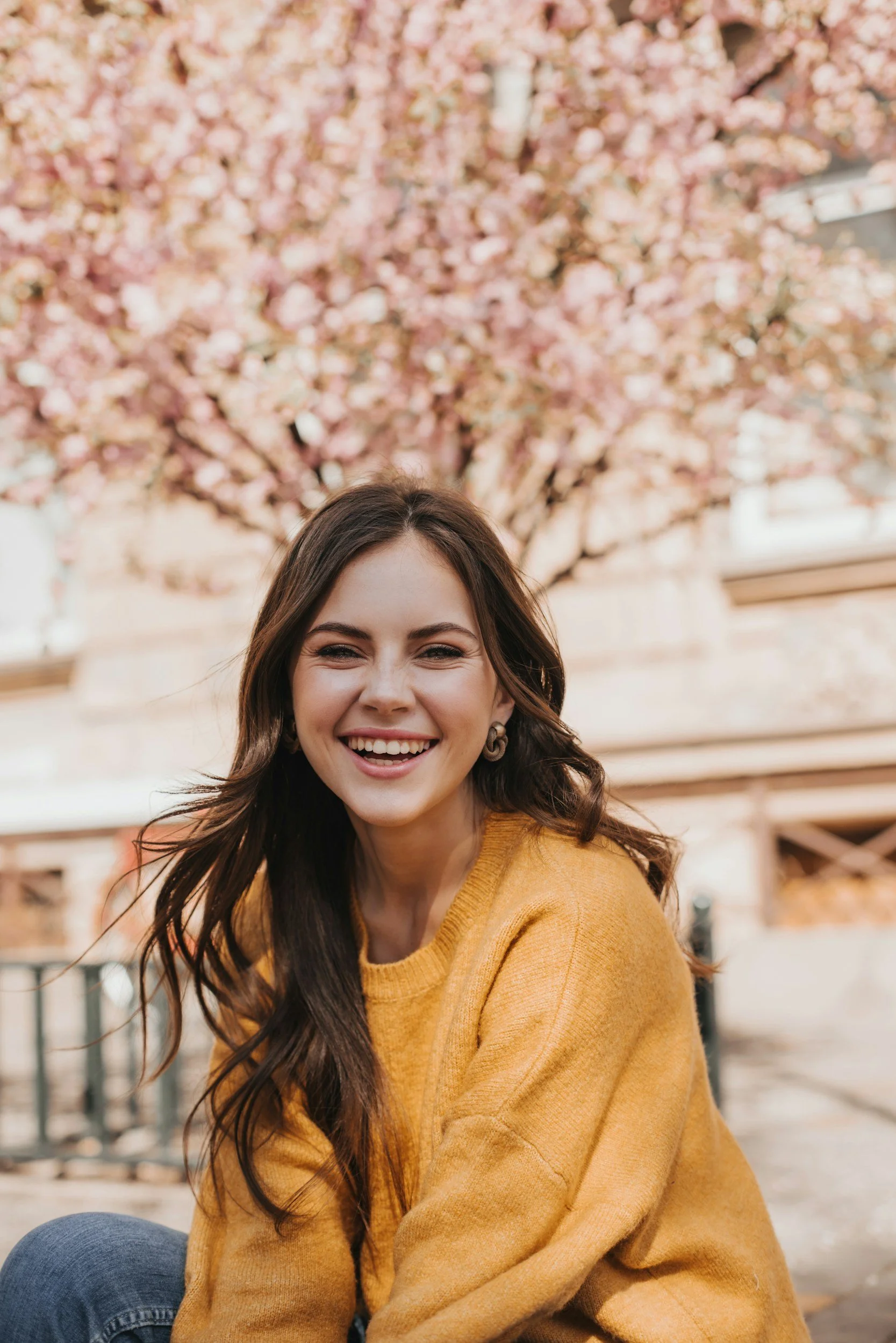 A smiling young woman with long brown hair wearing a yellow sweater, sitting outdoors in front of a blooming pink cherry blossom tree.