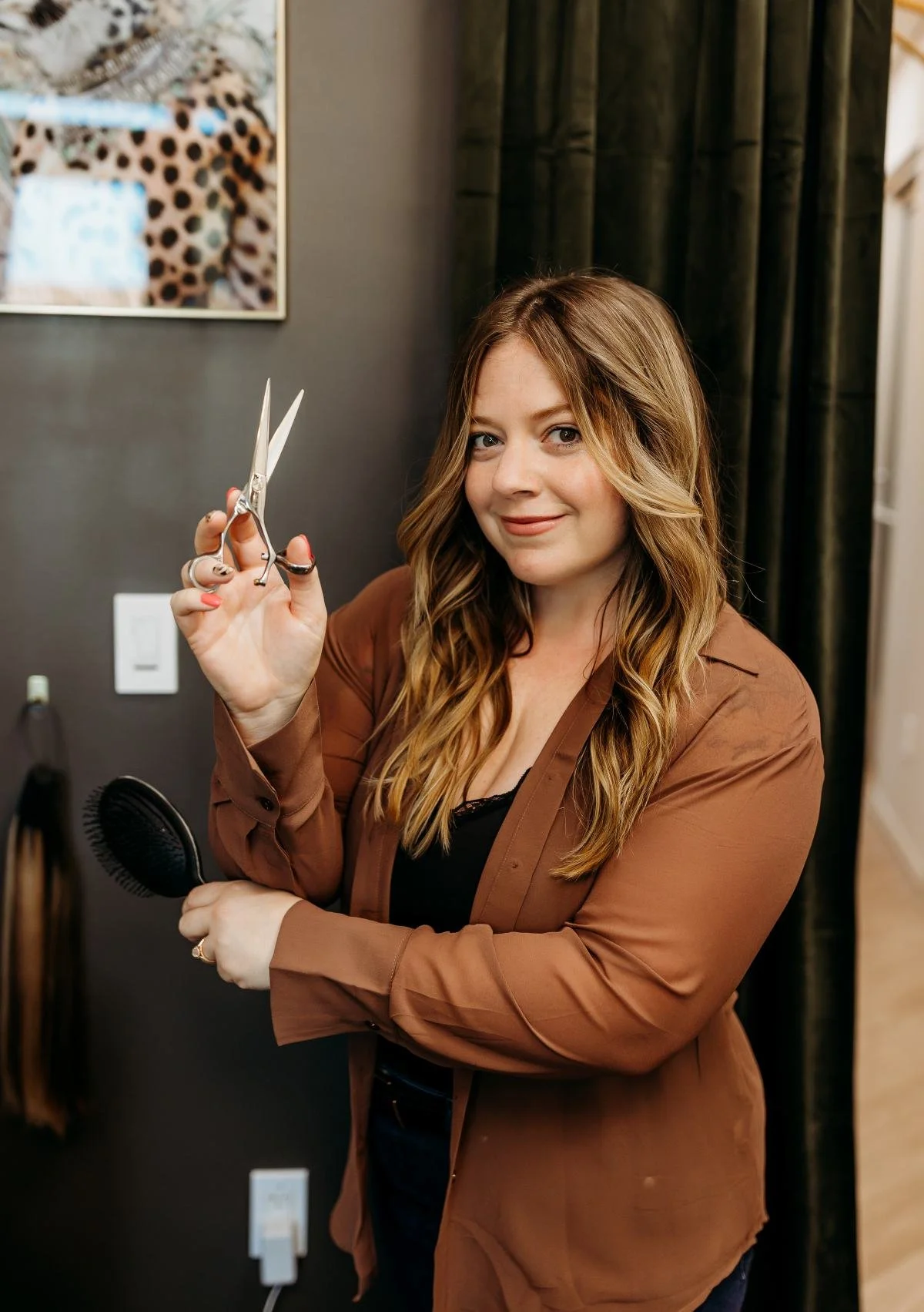 A woman with wavy, light brown hair holding a pair of scissors and a black hairbrush in a salon, smiling at the camera.