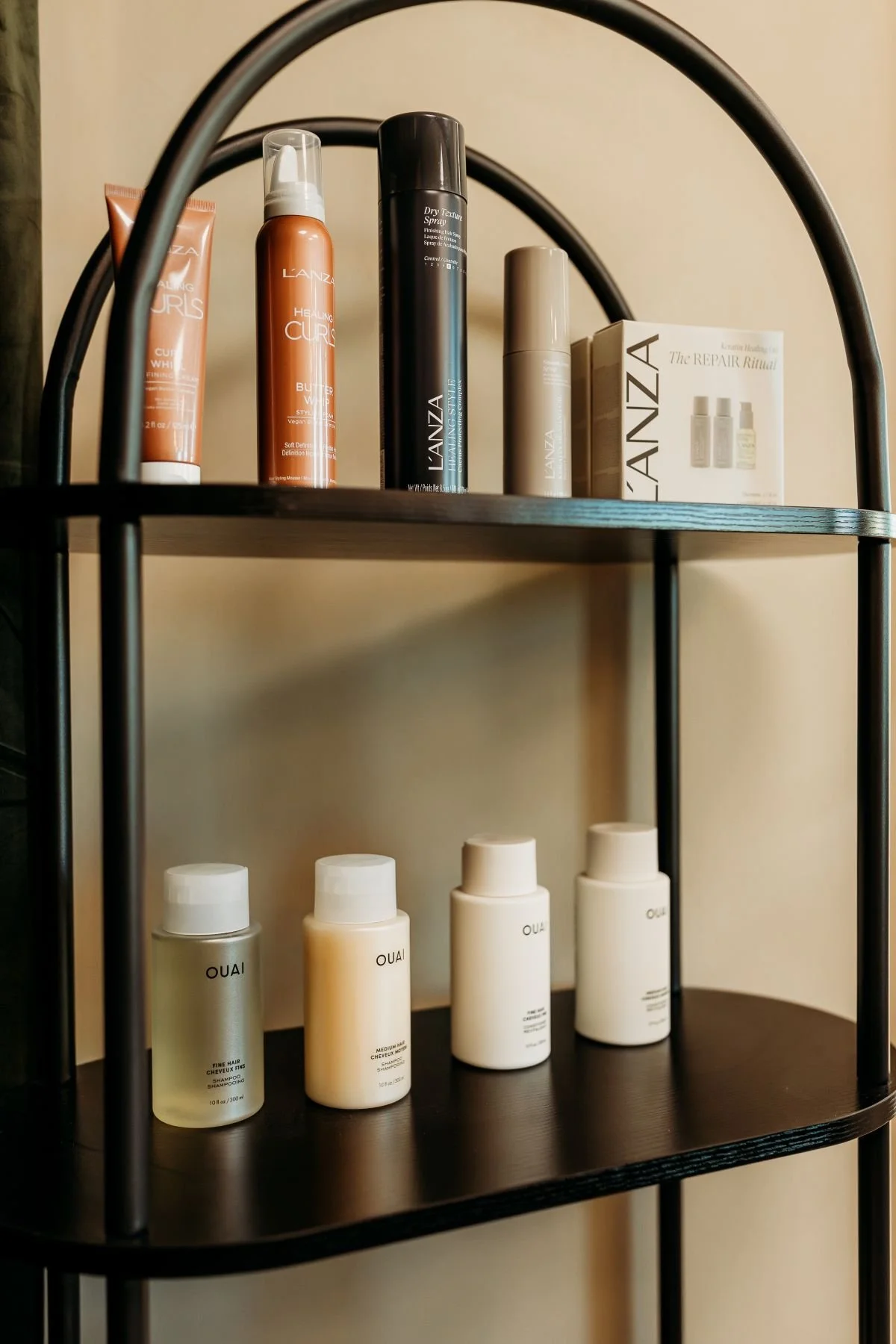 A black two-tiered shelving unit holding hair and scalp care products, including bottles of OUAI shampoo, conditioners, and other hair treatments.