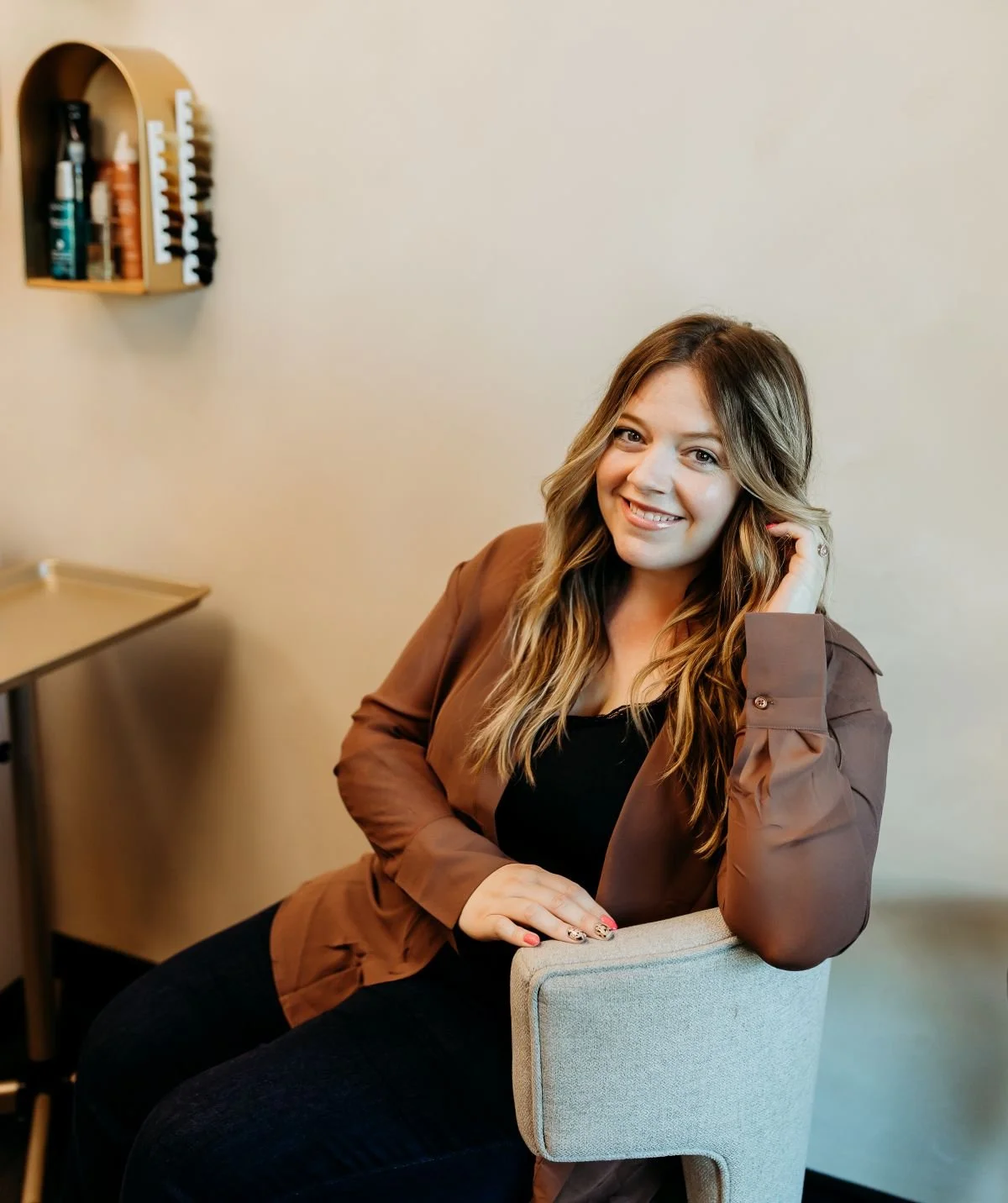 A woman smiling while sitting on a chair in a room with a beige wall and a shelf with bottles on it.