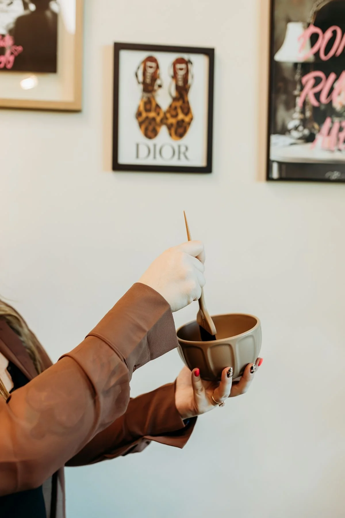 Person's hand holding a color bowl and stirring with a color brush, in cute salon with framed art and posters on the wall.