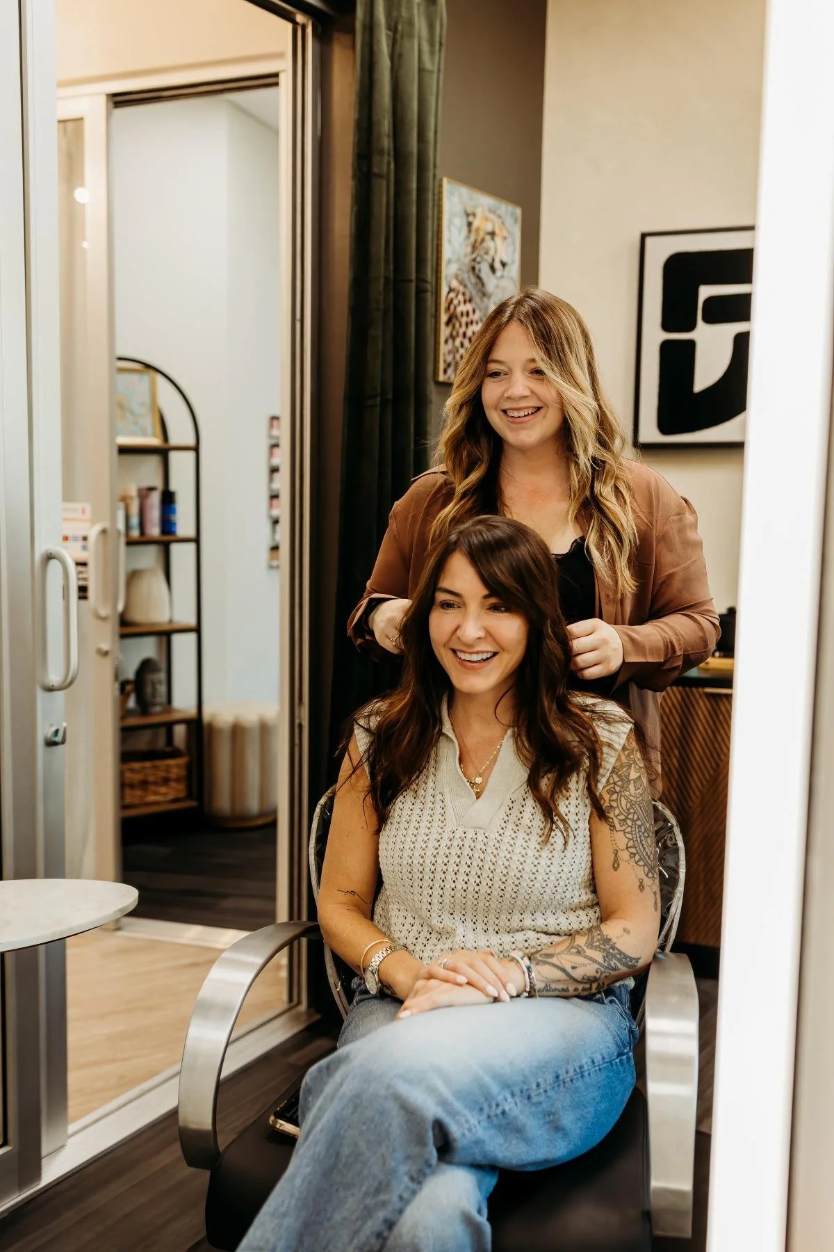 A woman with dark hair and tattoos sitting in a salon chair, smiling, while a hairstylist with long blond hair works on her.