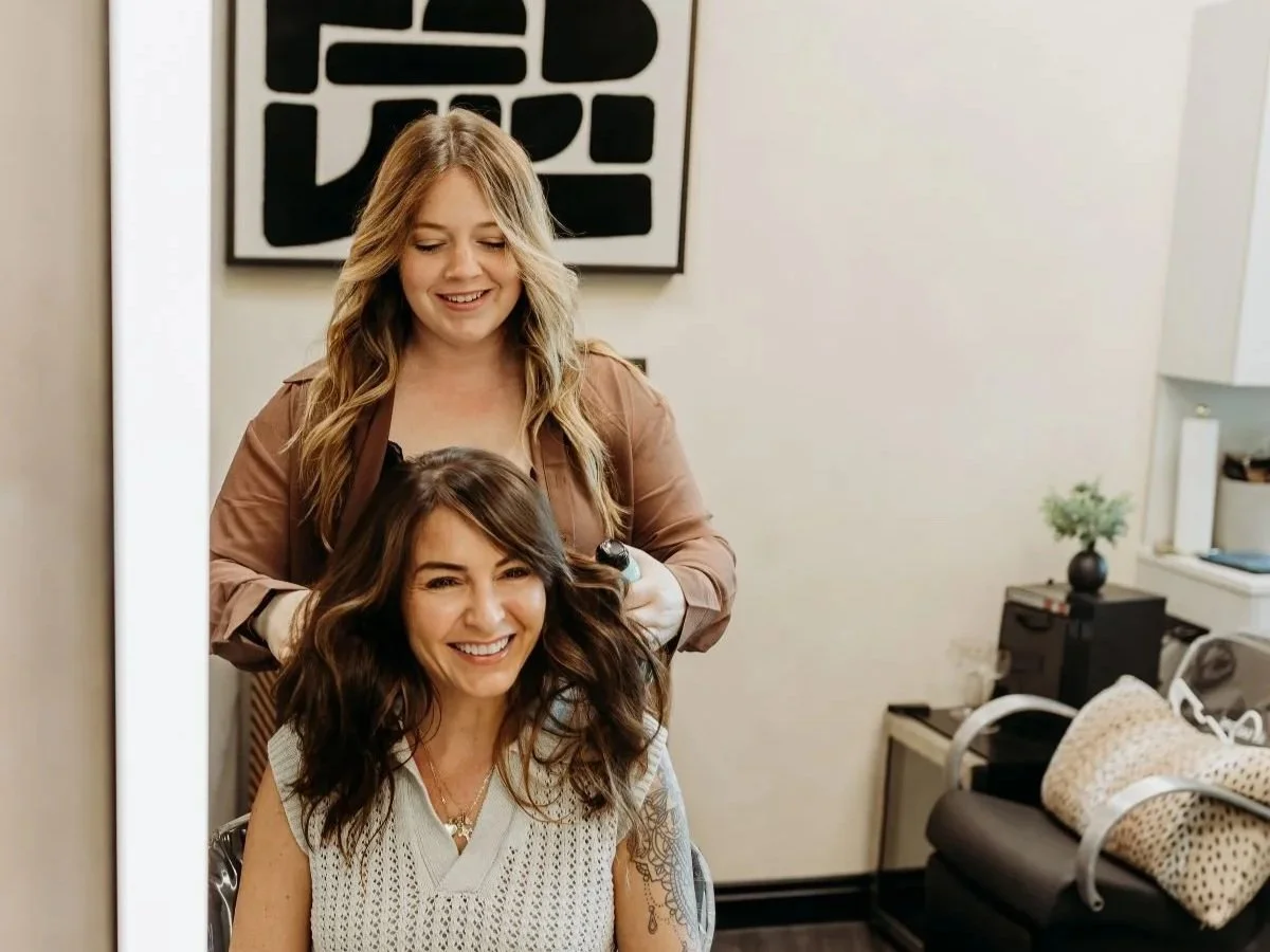 A hairstylist is styling a woman's hair in a salon, both are smiling, with a large black and white abstract art piece on the wall behind them.
