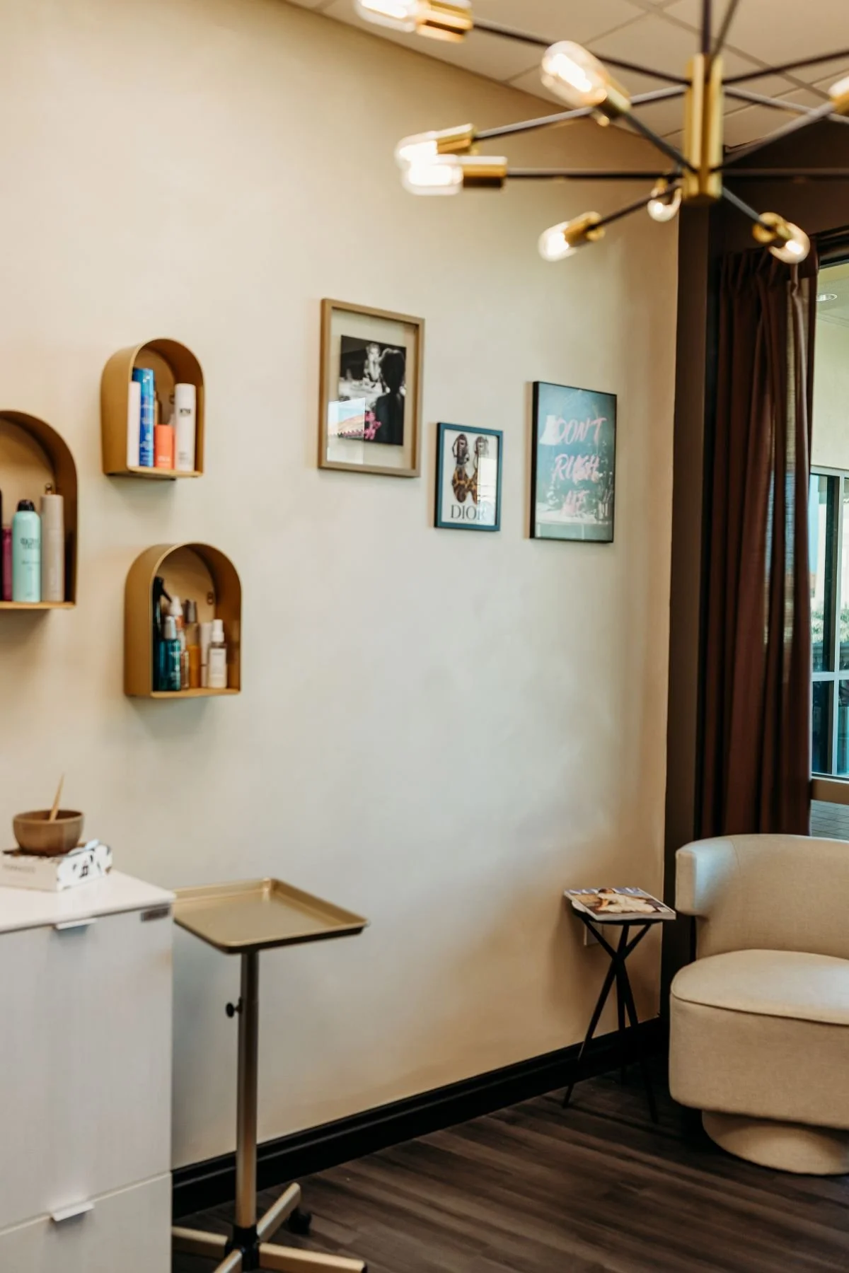 Interior of a room with a beige armchair, small black table with magazines, wall-mounted wooden shelves with skincare products, framed pictures on the wall, and a chandelier with multiple light bulbs. Curtains partly cover a window.
