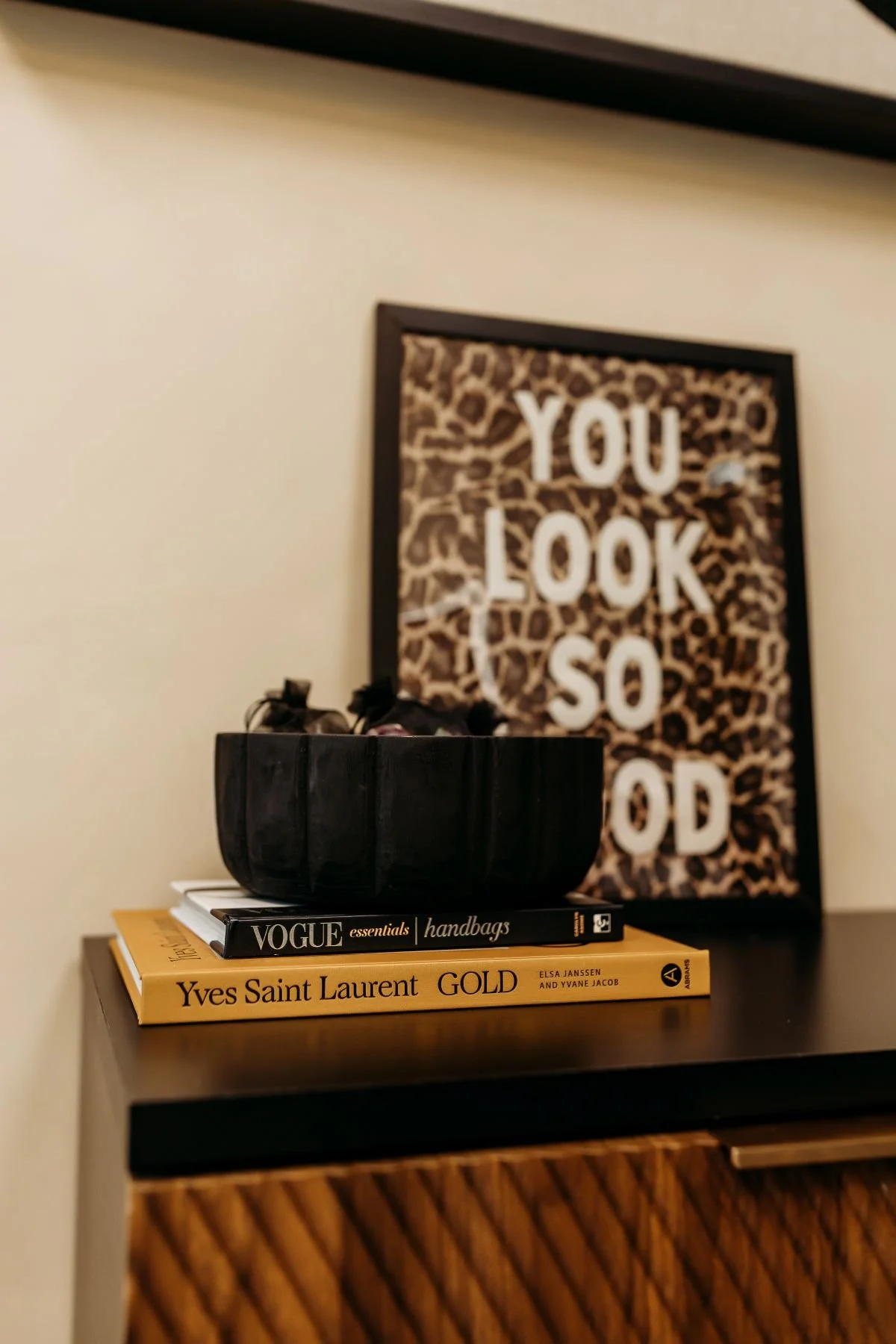 Stack of three books on a black surface, with a black bowl containing small objects, and a decorative framed sign with a leopard print background and white text that says "You Look So Good."
