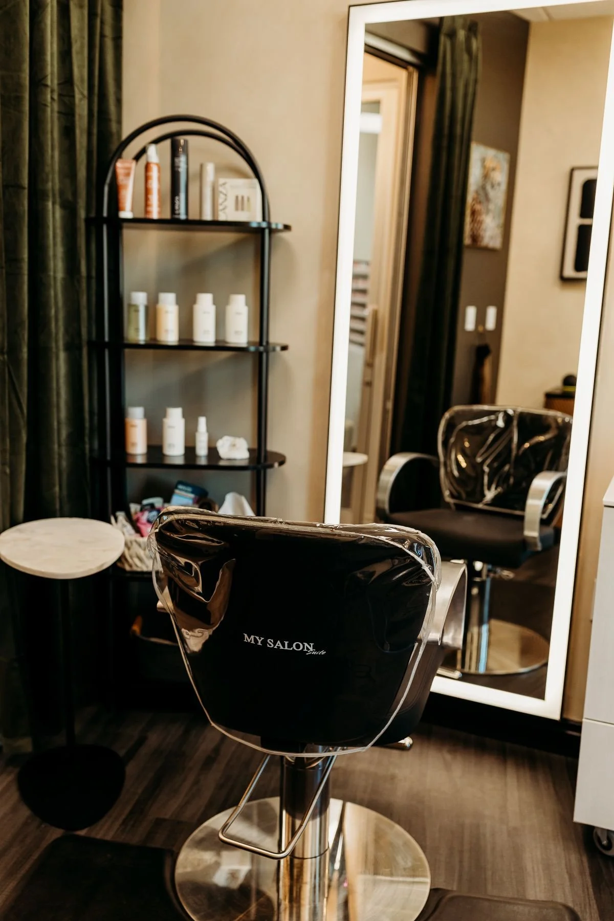 Empty black salon chair in front of a full-length mirror, with a small table and a plant shelf in the background.