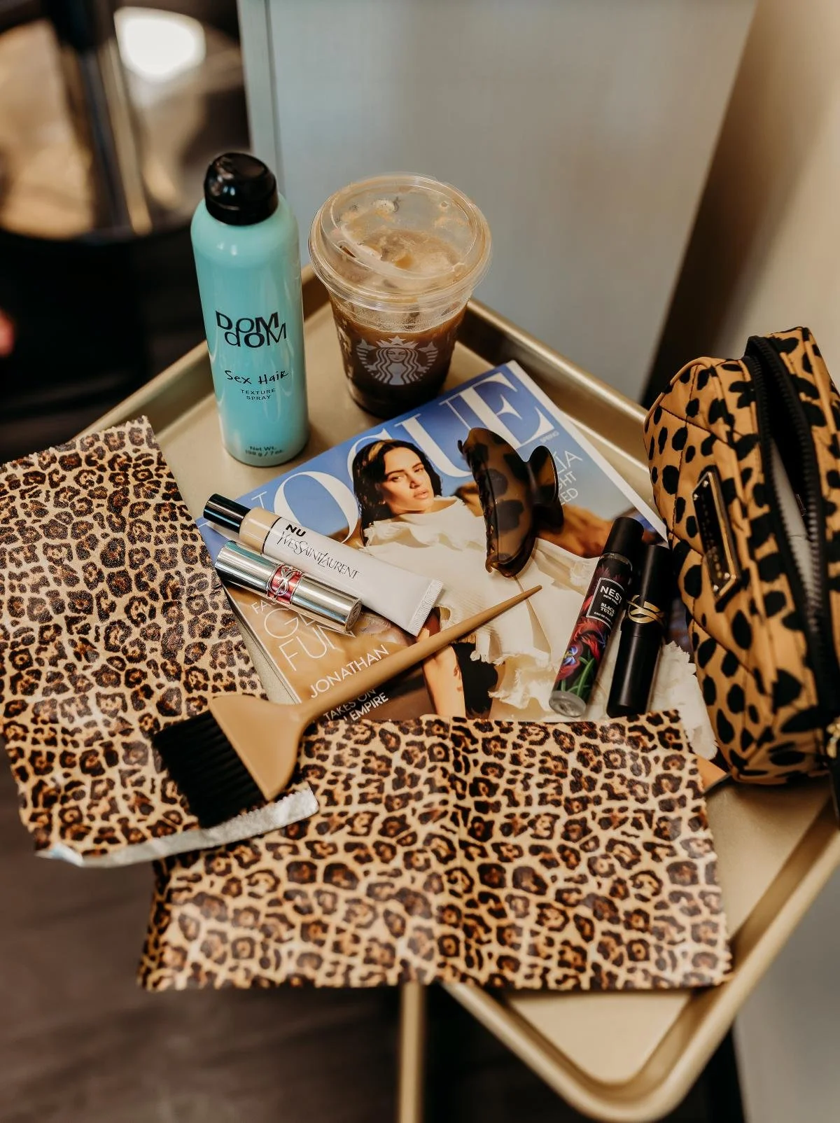 A table with various beauty and fashion items, including leopard print fabric, a bottle of dry shampoo, sunglasses, an iced coffee, a magazine, lipstick, a tube of skincare product, and a leopard print bag.