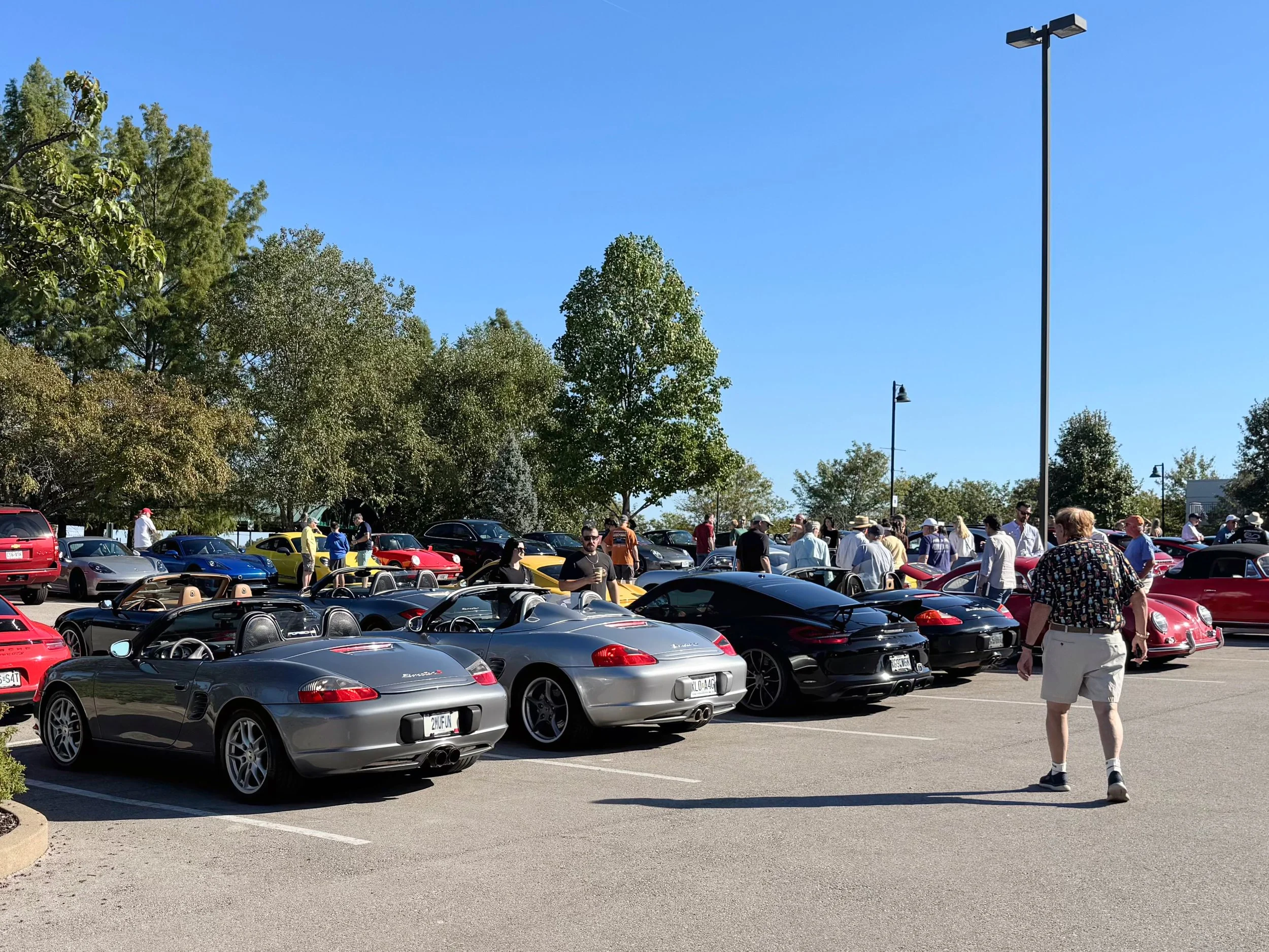 A car show in a parking lot with various sports cars and classic cars on display, and people walking around and inspecting the vehicles on a sunny day.