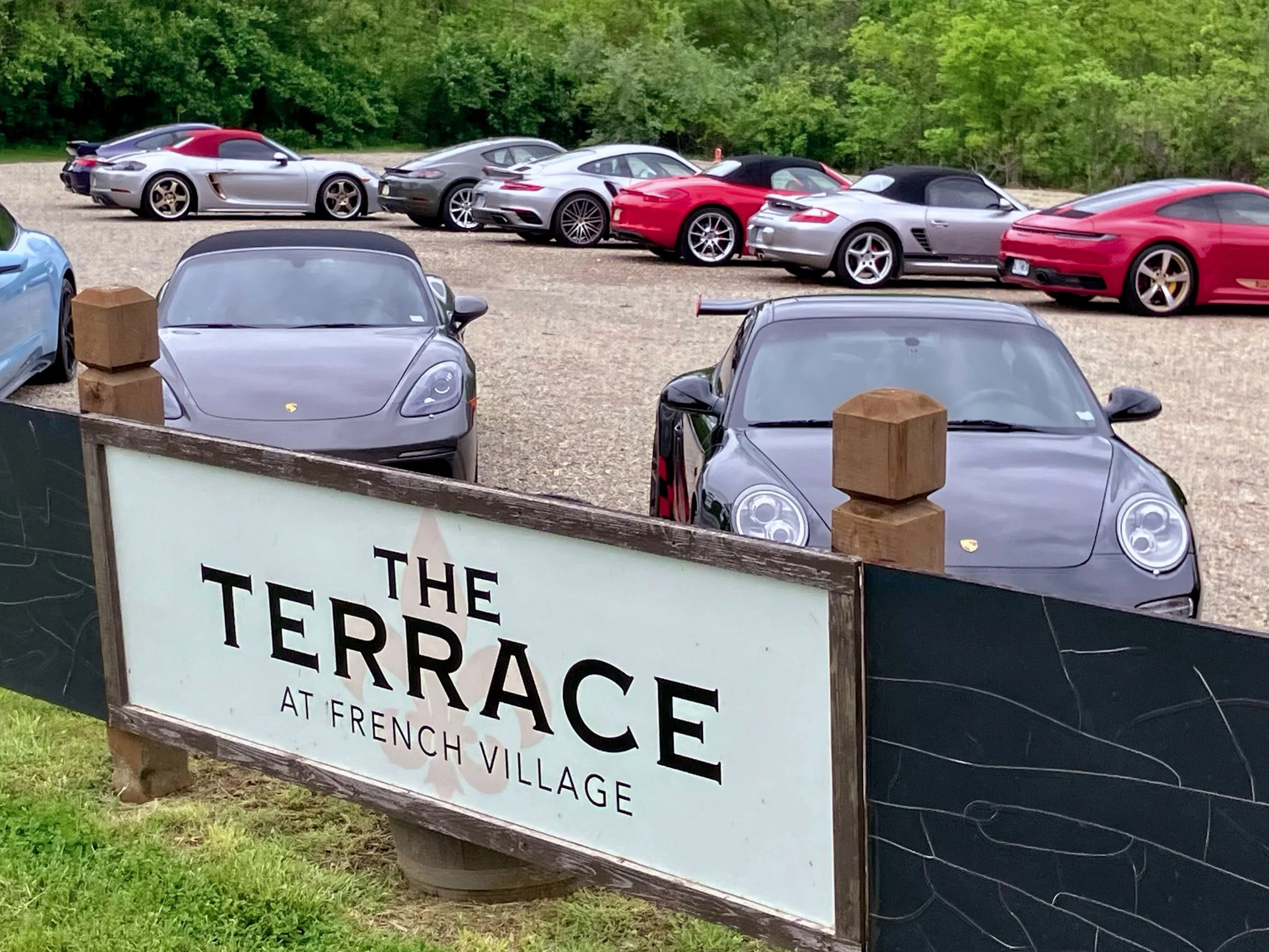 Several luxury sports cars parked behind a wooden sign that reads 'The Terrace at French Village' with a green, leafy background.