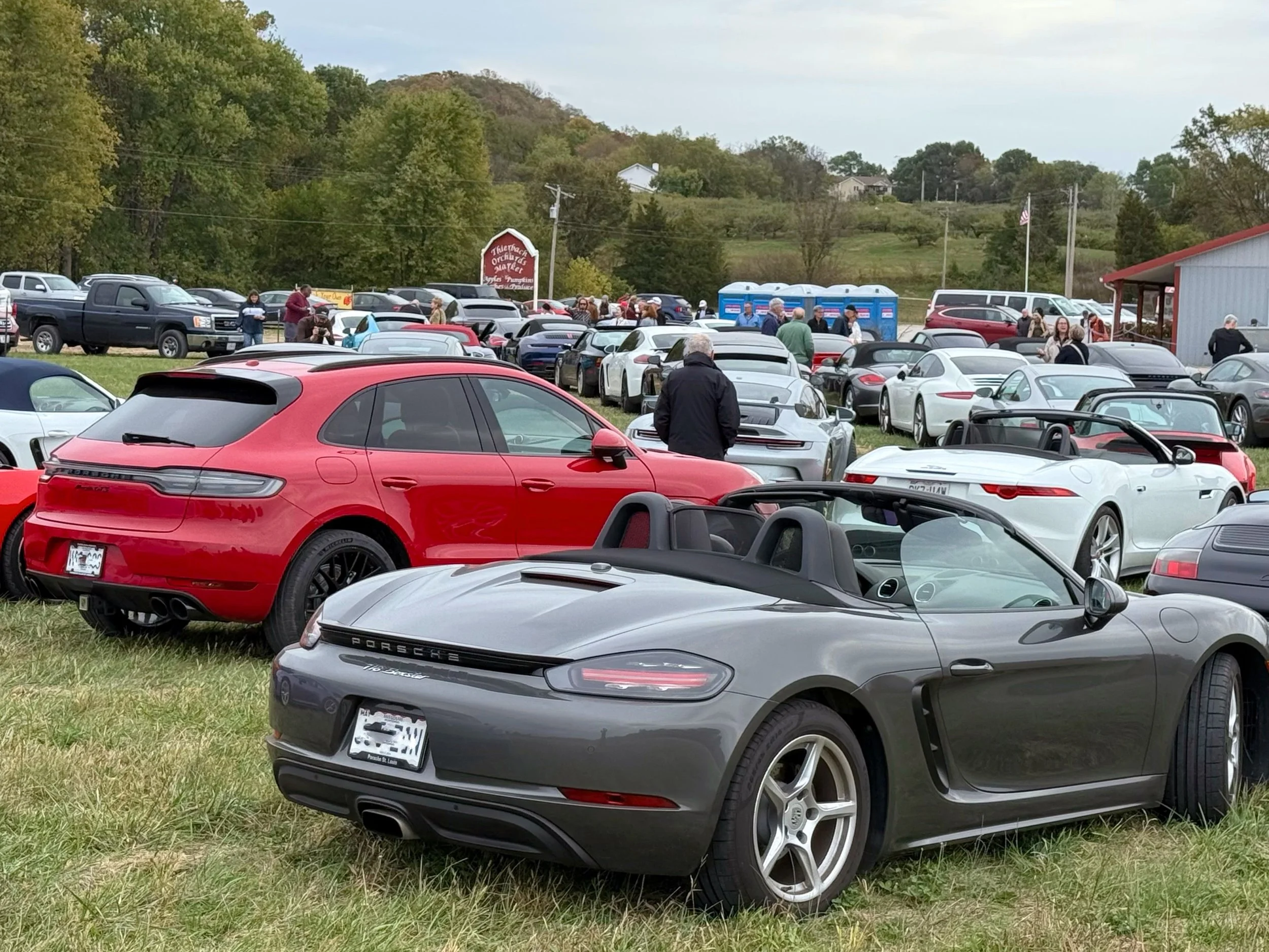 A collection of luxury cars, including gray Porsche and red Porsche SUVs, parked on a grassy field during an outdoor event with people walking around and a blue sky with clouds in the background.