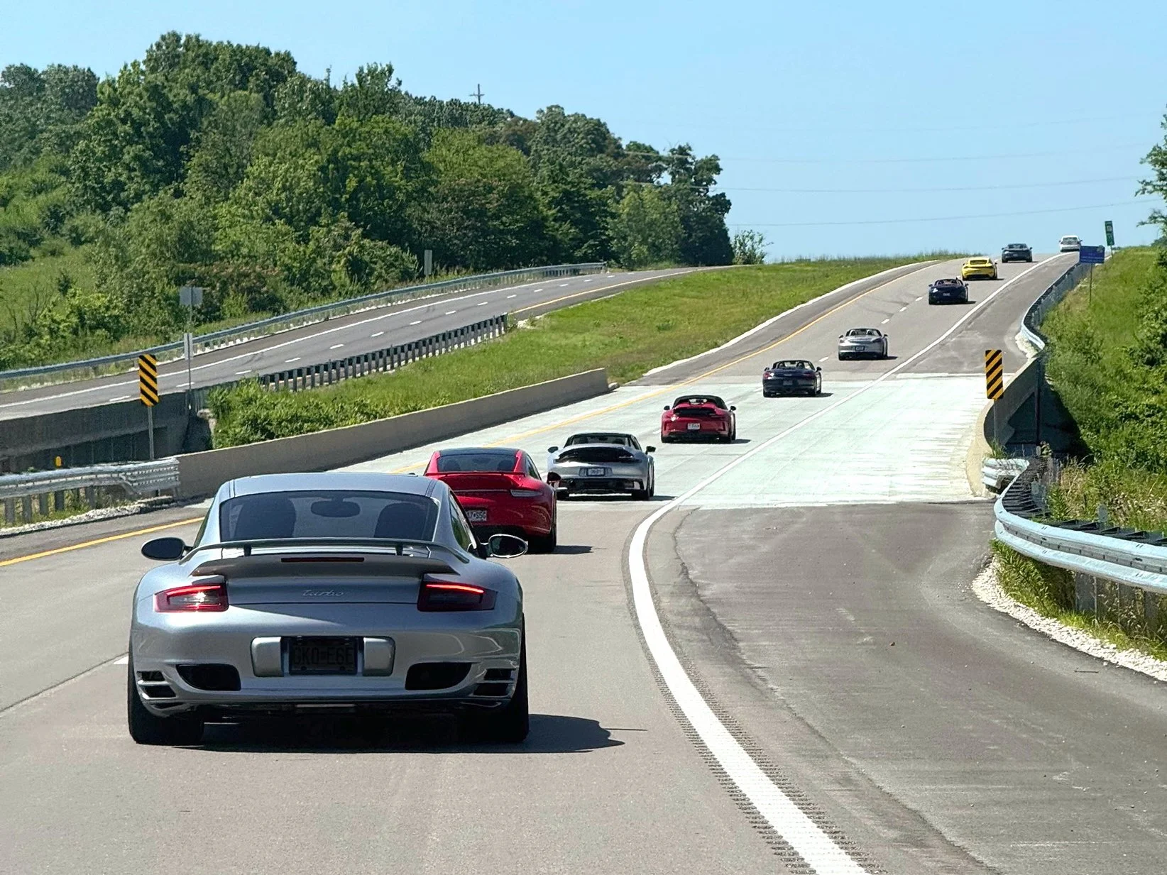 Multiple sports cars, including a silver Porsche 911 Turbo, traveling on a two-lane highway with a scenic, hilly landscape and green trees on a sunny day.