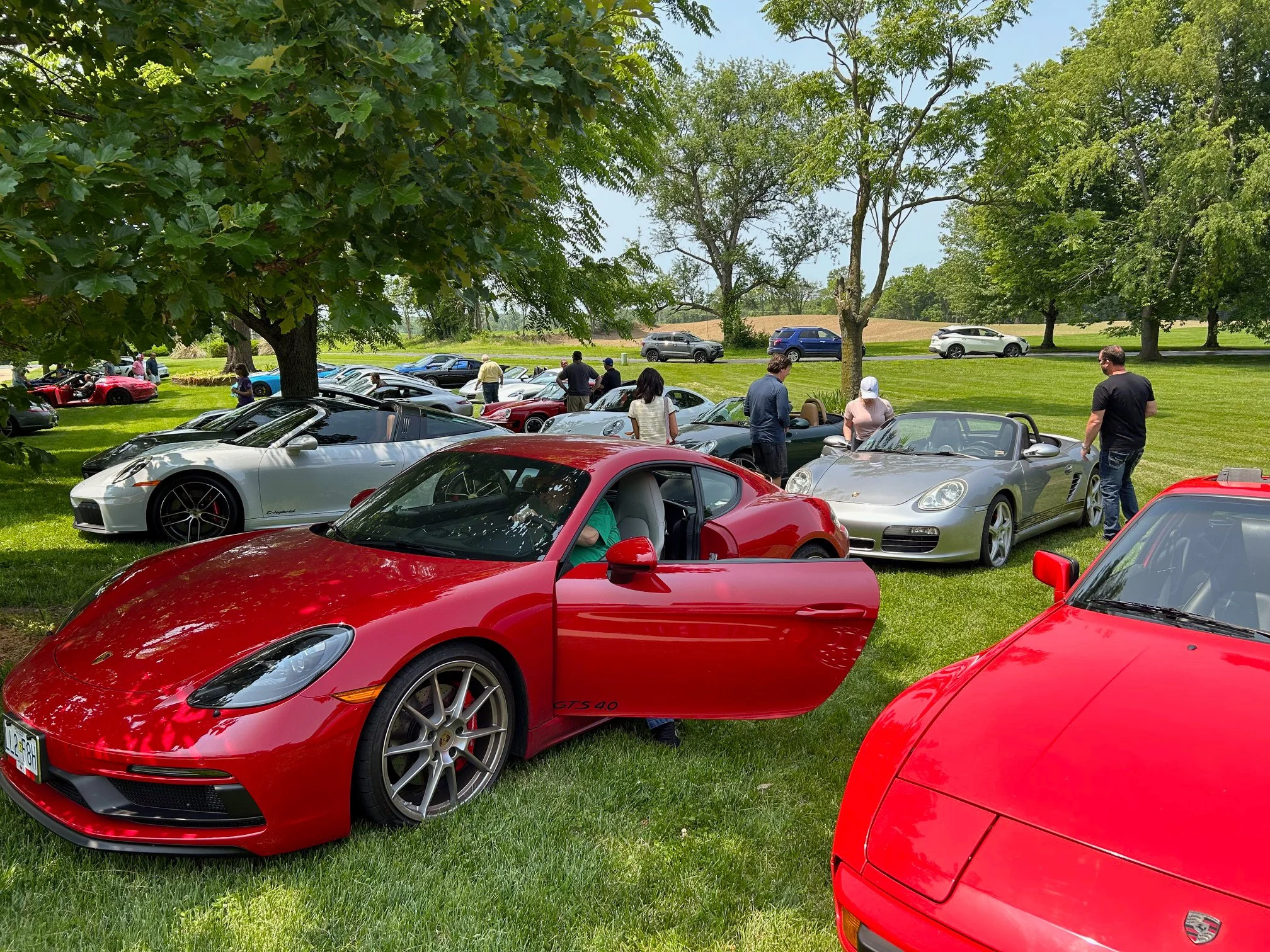 A gathering of luxury cars, including a red Porsche 718, silver Porsche Boxster, red Porsche 911 GTS 40, and a silver Corvette, parked on a grassy area under trees, with people admiring and examining the vehicles on a sunny day.
