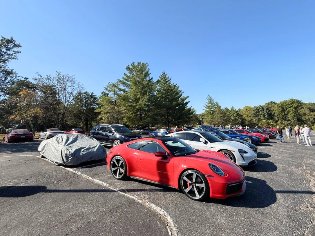 A red sports car parked in a lot among various other cars under a blue sky with trees in the background.