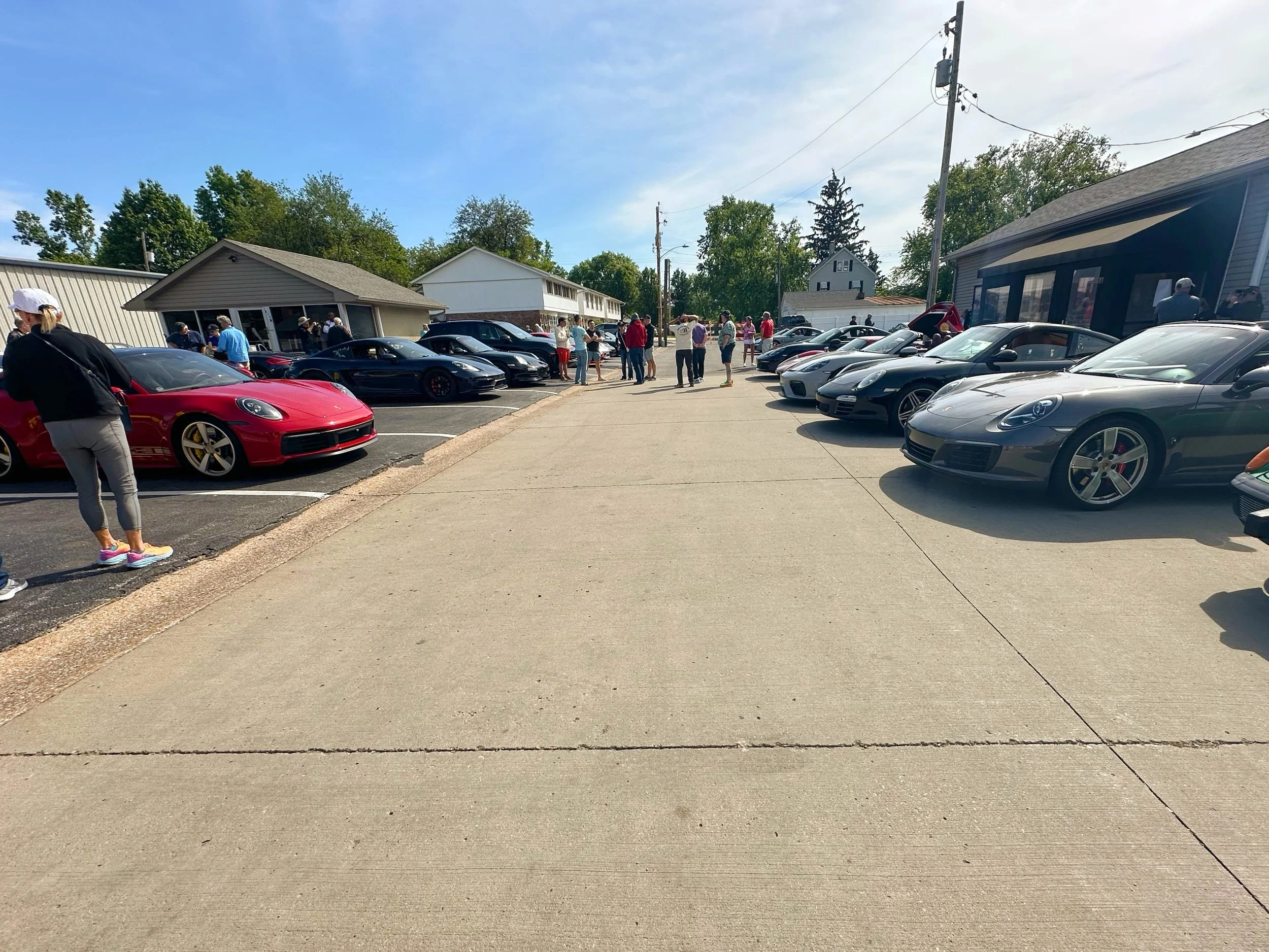 A gathering of people around luxury sports cars parked on a lot on a sunny day, with a group of people standing near the cars and some buildings in the background.