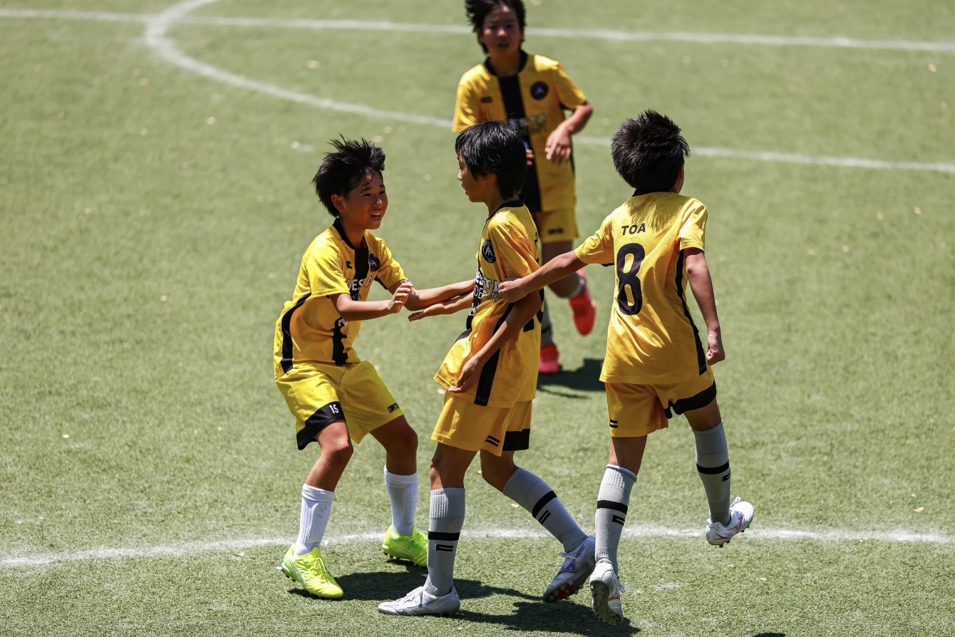 Group of young boys in yellow soccer uniforms celebrating and hugging on a grassy field during a game.