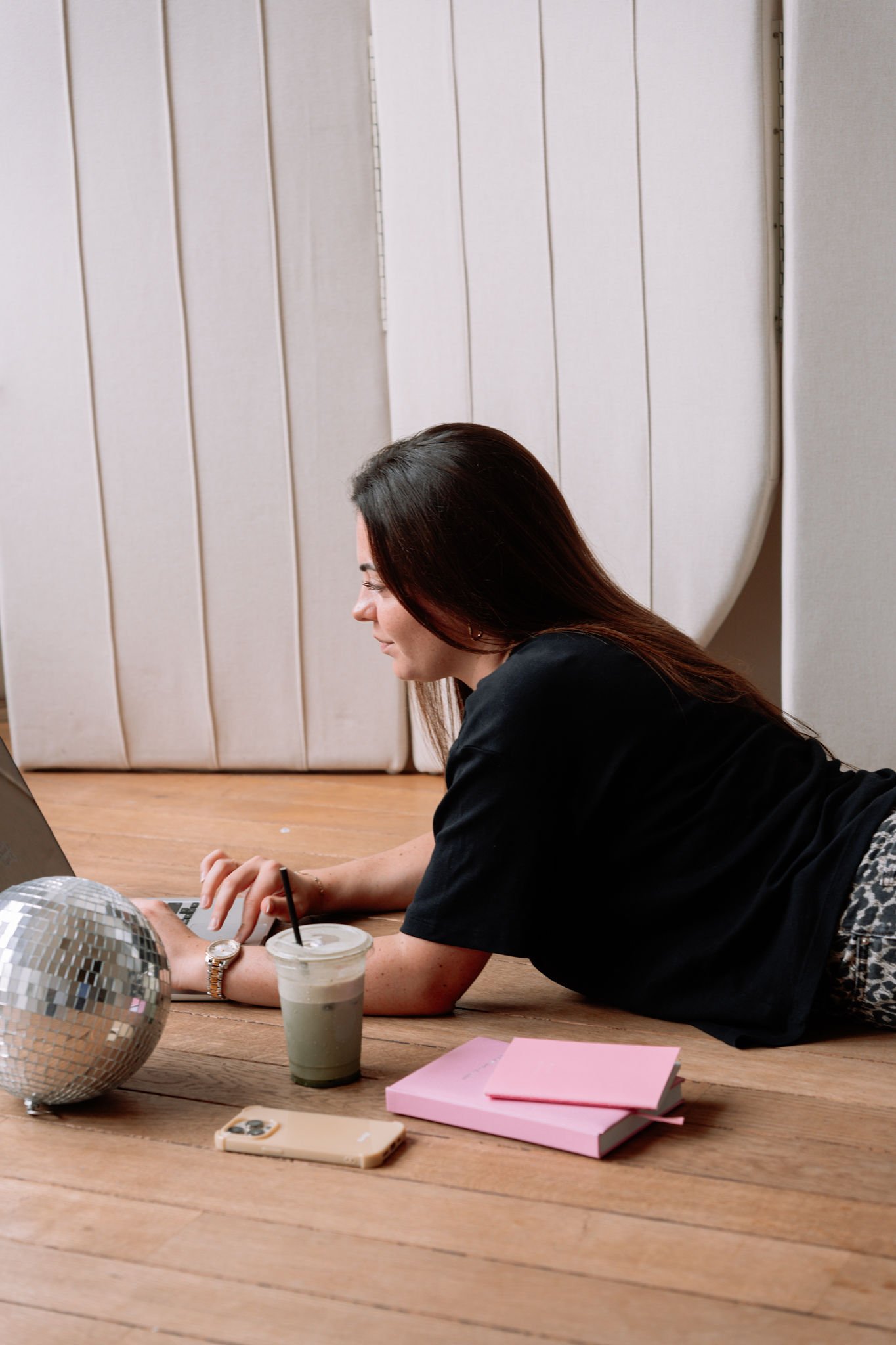 Une femme allongée sur un sol en bois, utilisant un ordinateur portable, avec un verre à boisson, deux cahiers roses, un téléphone portable en couverture beige, une boule disco argentée et d'autres objets à proximité.