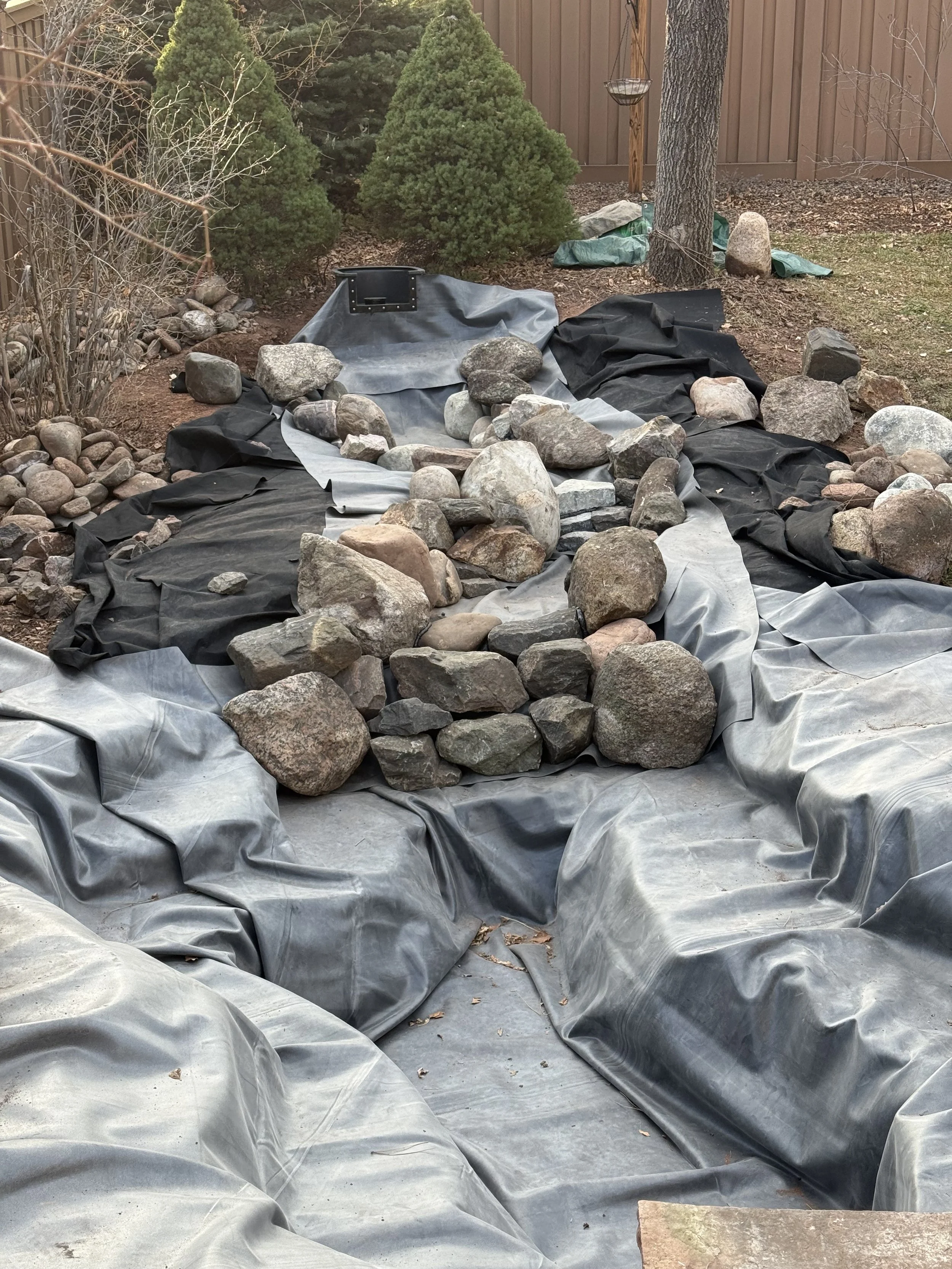 A backyard pond under construction with a black pond liner and rocks placed inside, surrounded by soil, trees, and a wooden fence in the background.