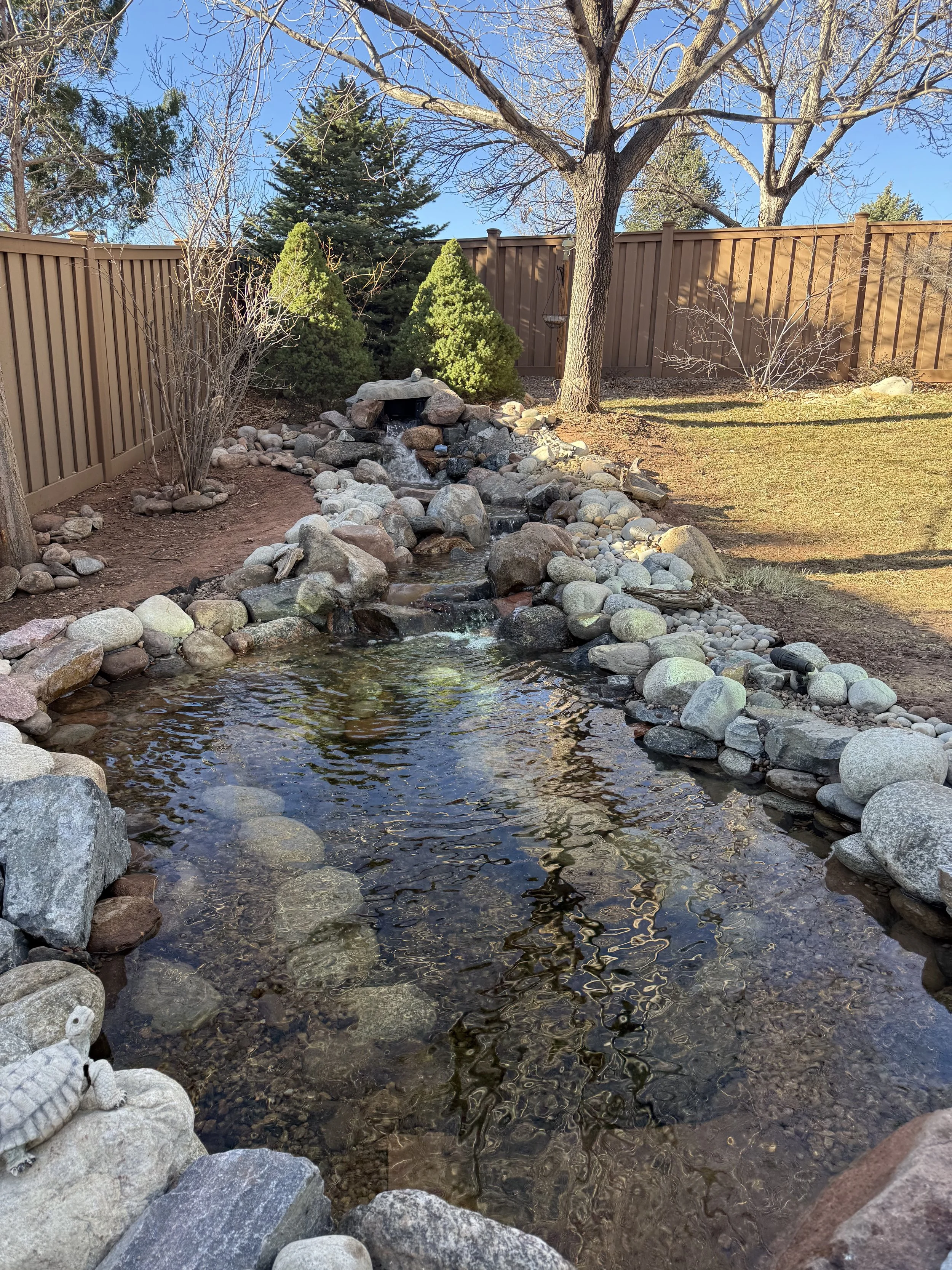 Backyard garden with a small rock-lined stream, a small rock waterfall, a leafless tree, evergreen bushes, a wooden fence, and a partly cloudy sky.