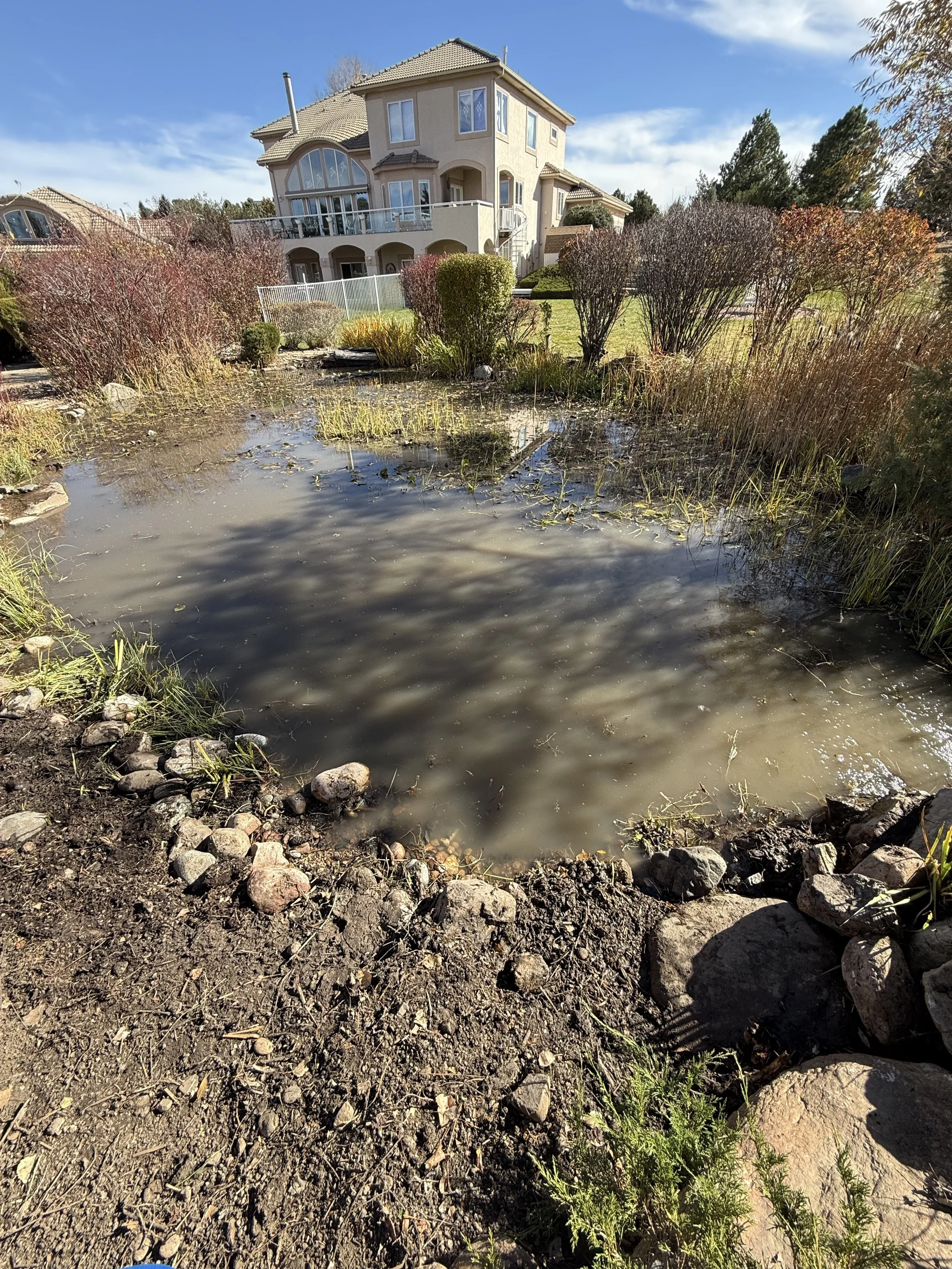 A pond with cloudy water surrounded by rocks and plants in front of a large beige house with multiple windows and a balcony on a sunny day.