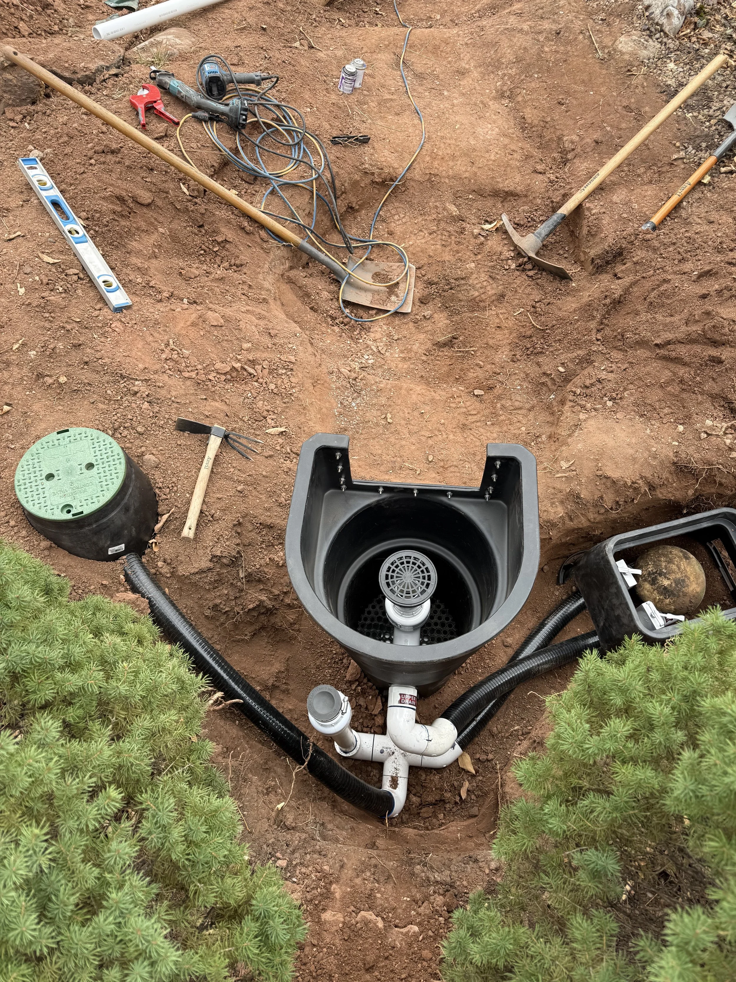 A partially installed underground drainage system with pipes, a sump basin, and surrounding tools at a construction site.
