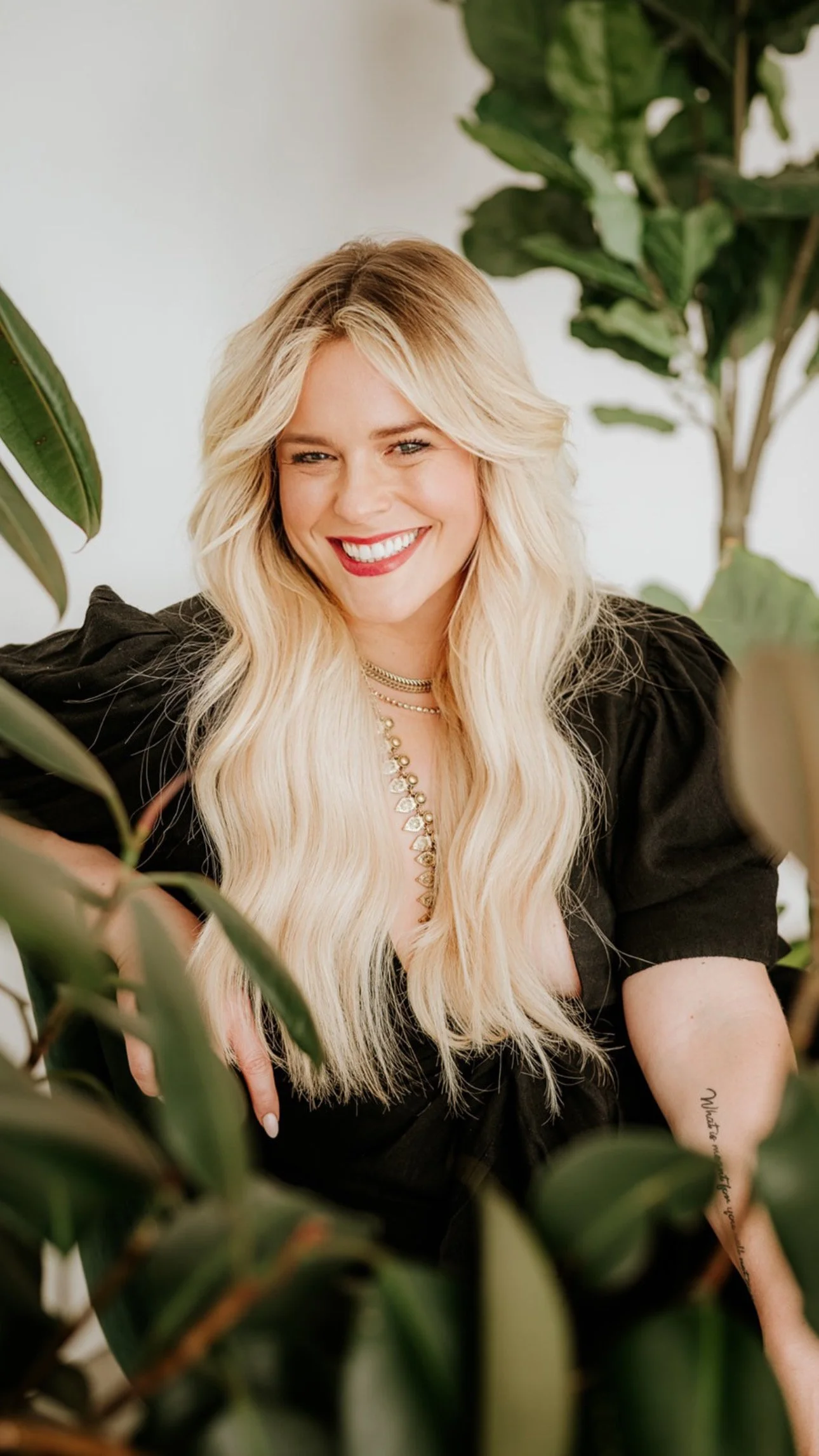 A woman with long blond hair, smiling, wearing a black top, and jewelry, surrounded by green plants.