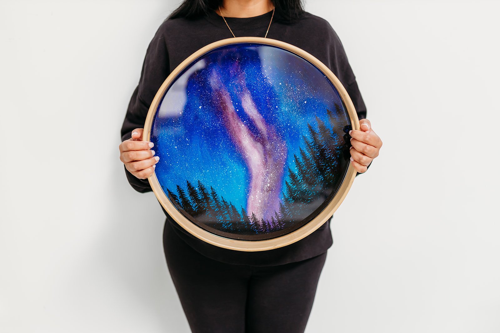 Person holding a circular wooden frame featuring a painting of a starry sky with a galaxy and silhouetted trees at the bottom.