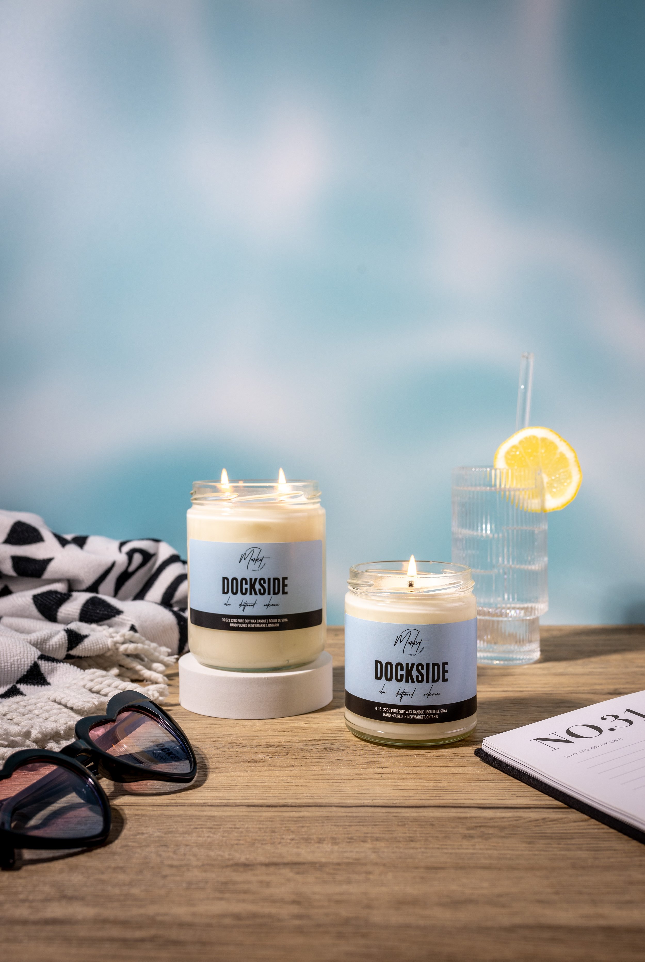 Two Dockside scented candles on a wooden surface next to a glass of water with a lemon slice, heart-shaped sunglasses, and a notebook. A patterned towel is draped nearby.