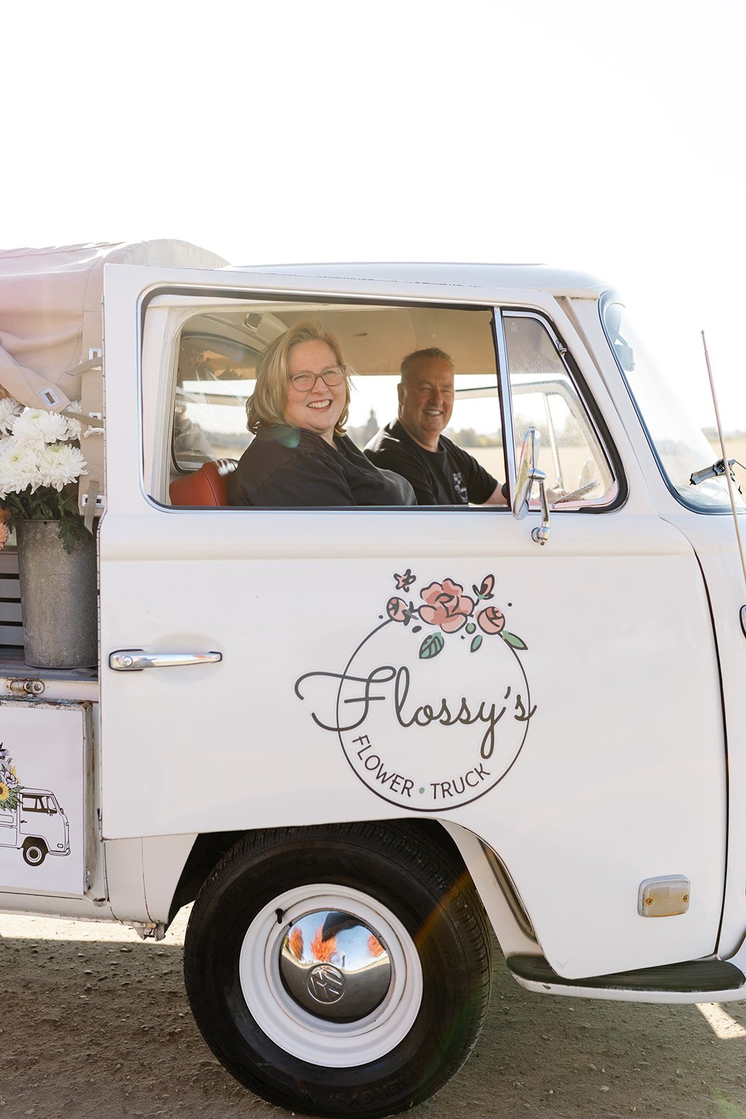 Two people sitting inside a vintage white flower truck with the logo "Flossy's Flower Truck" adorned with floral design. The truck has flowers in the back.