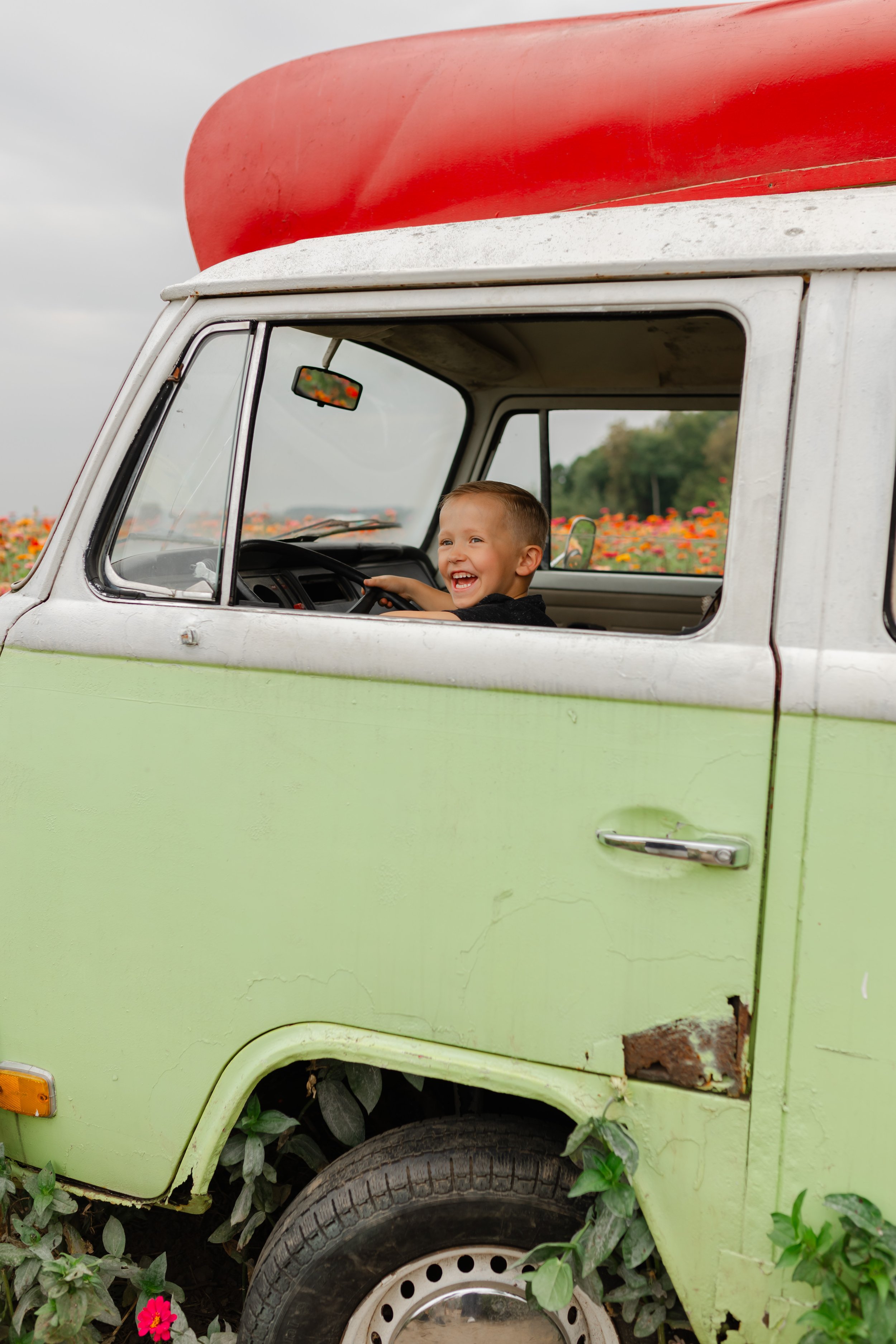 Child smiling inside an old green van with a red roof against a background of flowers.
